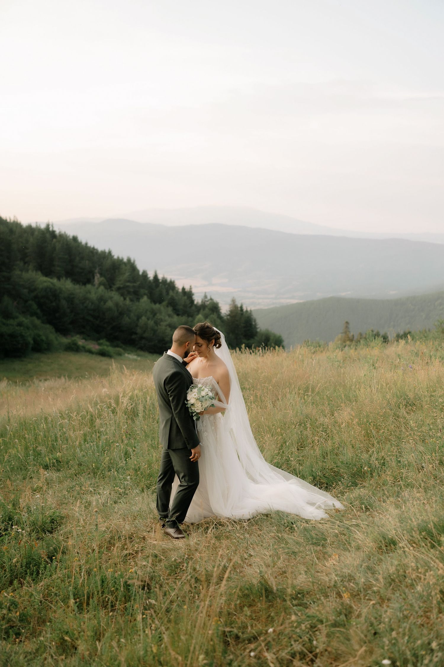 A couple in wedding attire share an intimate moment in a scenic mountain meadow at sunset.