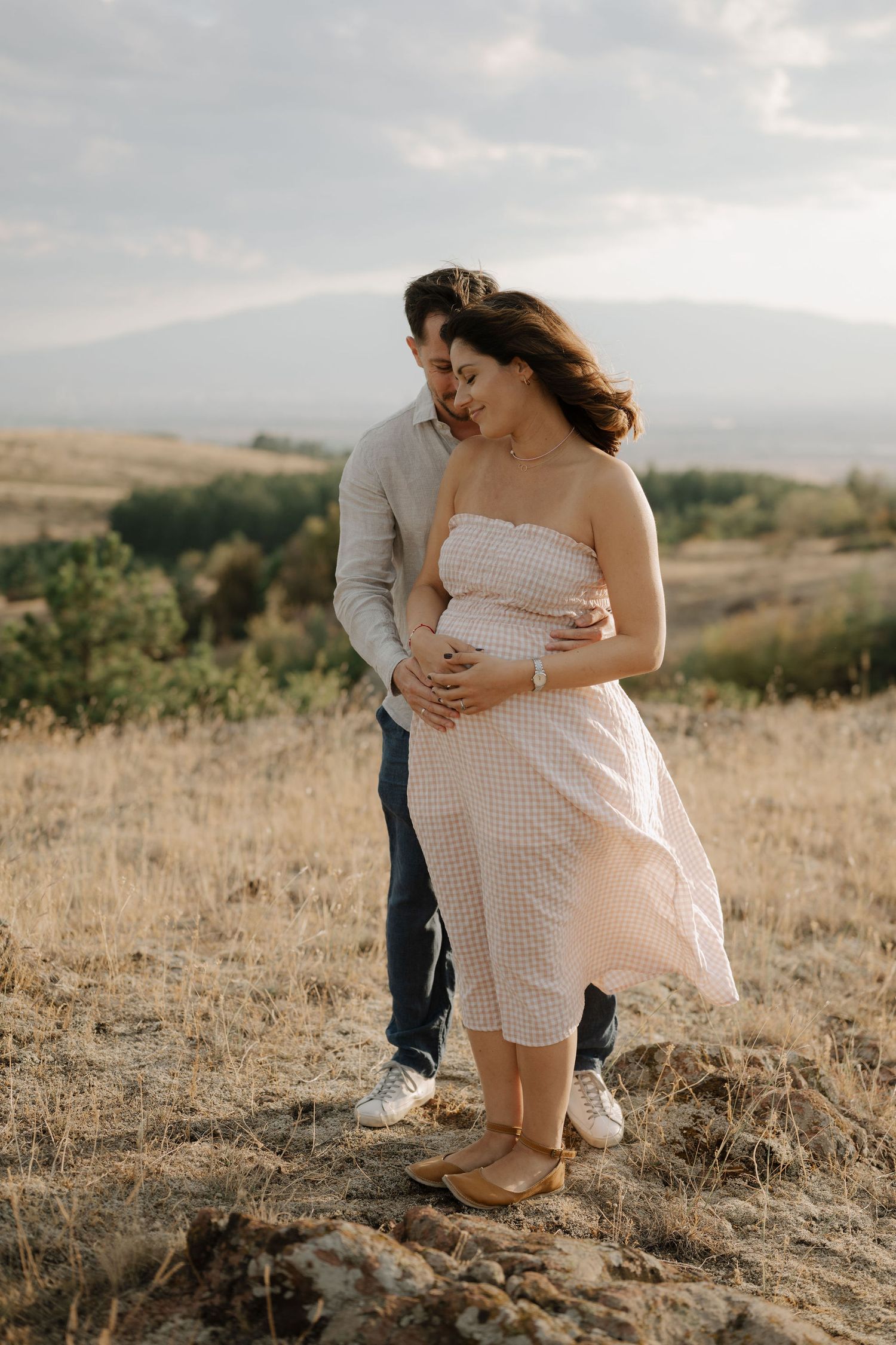 Couple embraces in a sun-drenched field with rolling hills in the background during golden hour.
