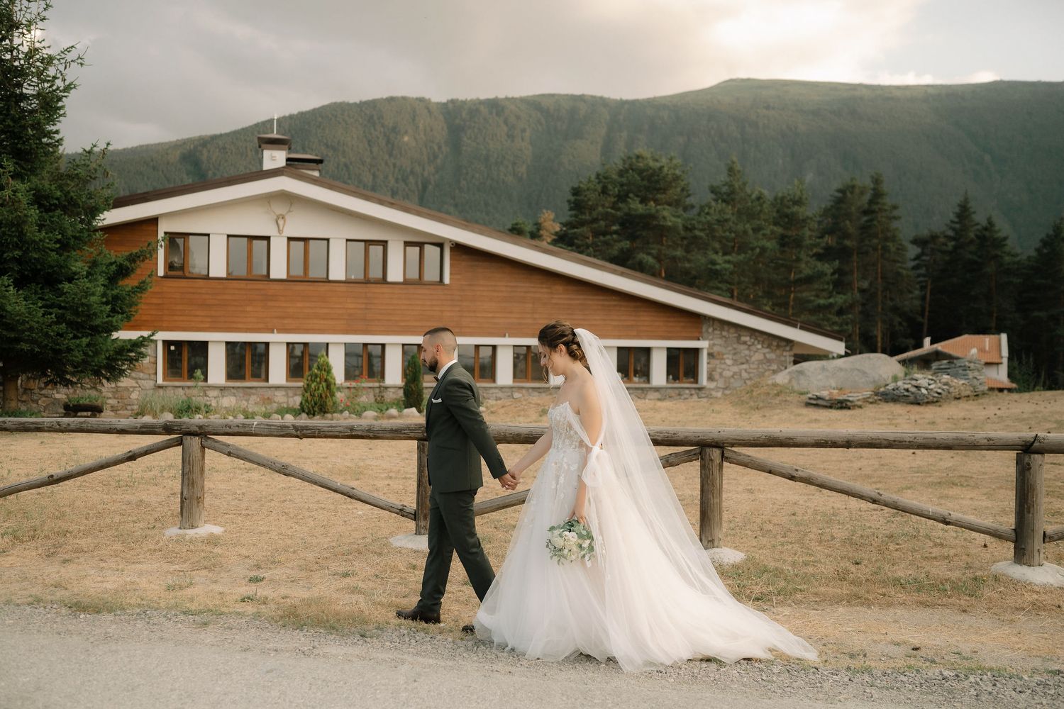 Panoramic view of wedding couple near mountain lodge with cloudy skies and evergreen trees in background.