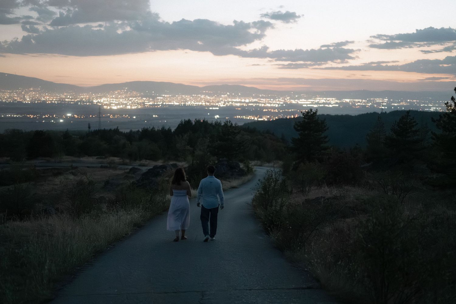 A series of silhouettes walking on a scenic trail at sunset with mountains and city lights in the background.