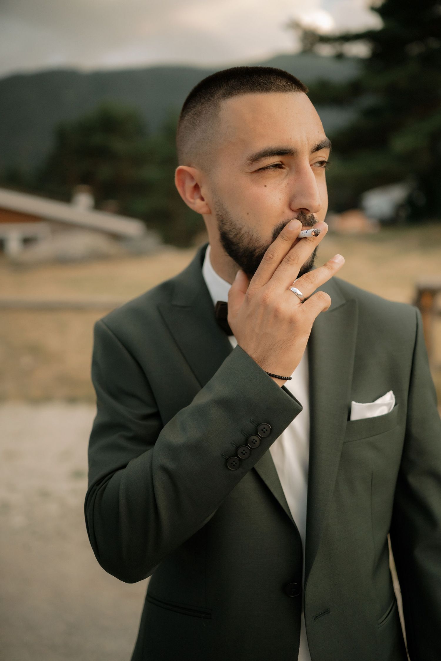 A person in a dark green suit and white pocket square smokes a cigarette while looking contemplatively into the distance.
