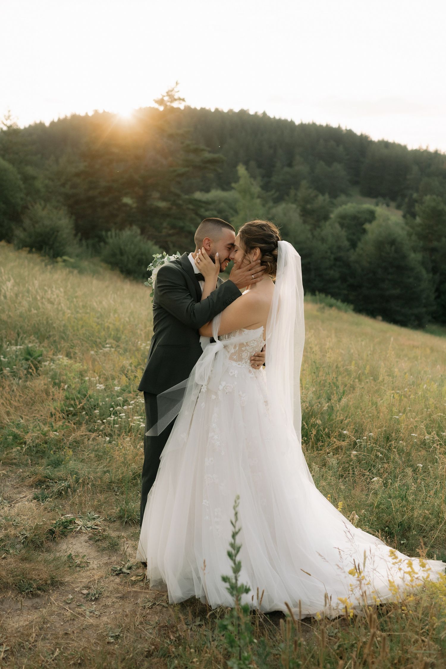 A romantic wedding couple shares intimate moments at sunset in a golden meadow with flowing white dress and veil.