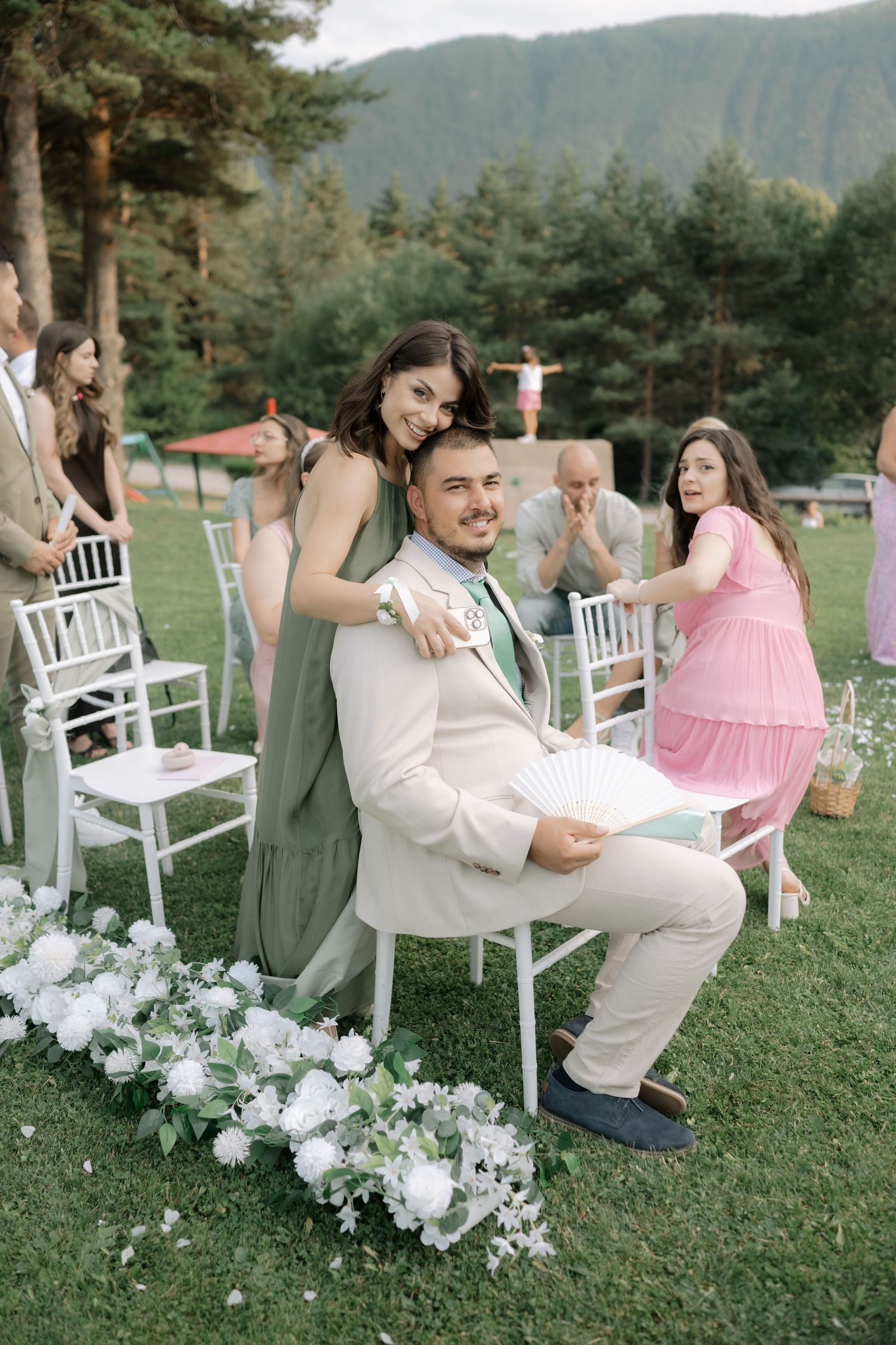 Wedding party members playfully pose with white chairs during an outdoor ceremony setup in a mountain setting.