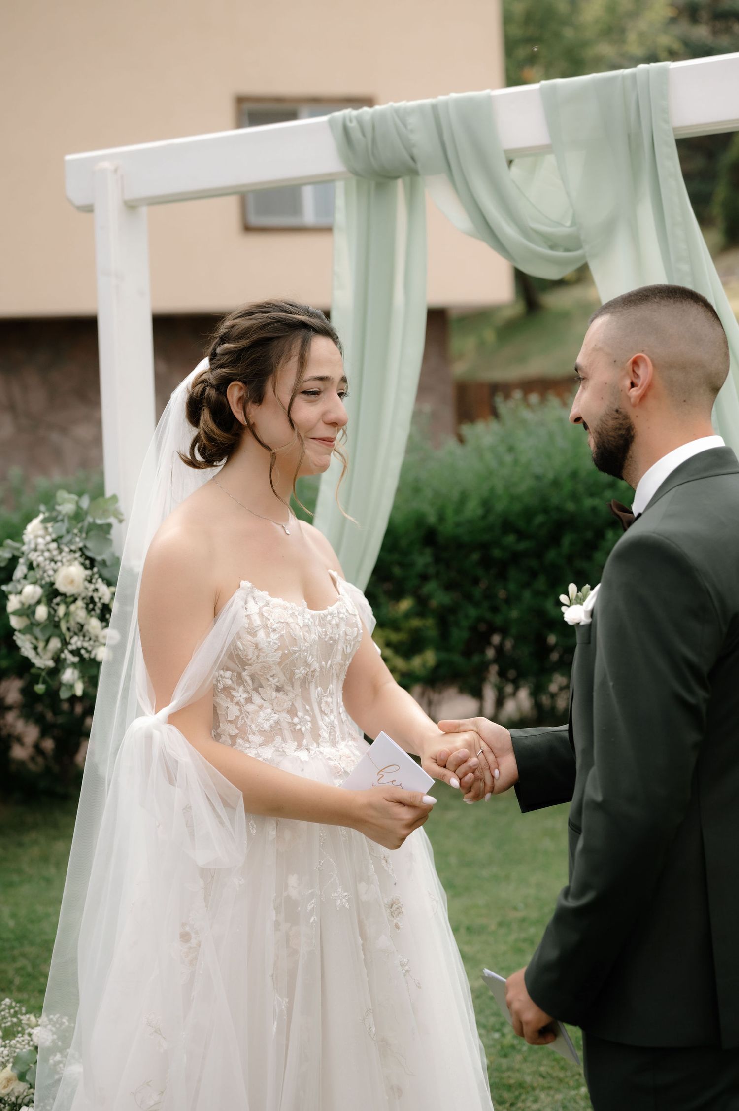 An intimate wedding ceremony moment under a white arch decorated with sage green fabric and white flowers.