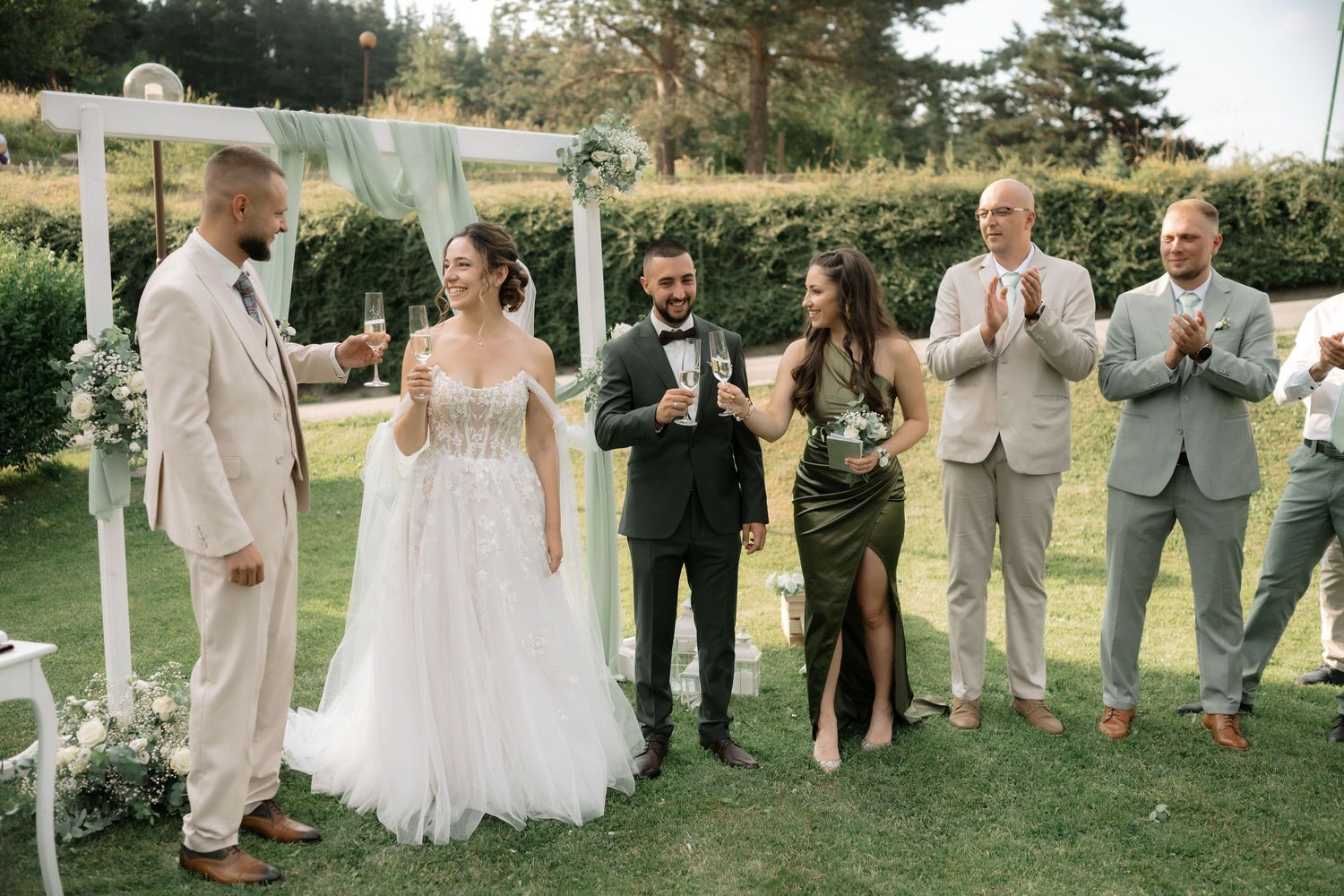 Wedding party stands on grass lawn during outdoor ceremony with white fabric backdrop and floral decorations.