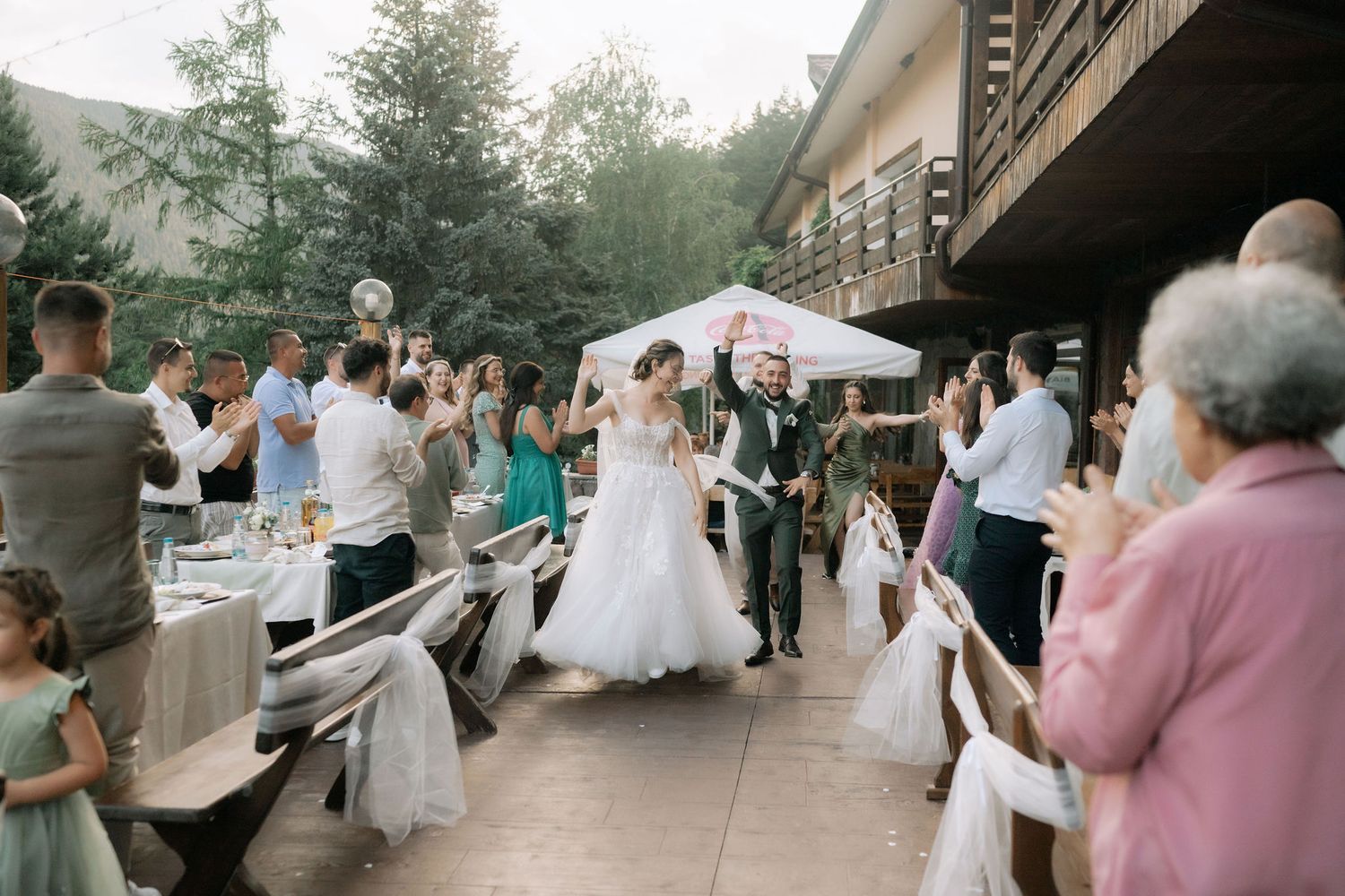 Wedding guests dance and celebrate at an outdoor reception venue surrounded by trees and string lights.
