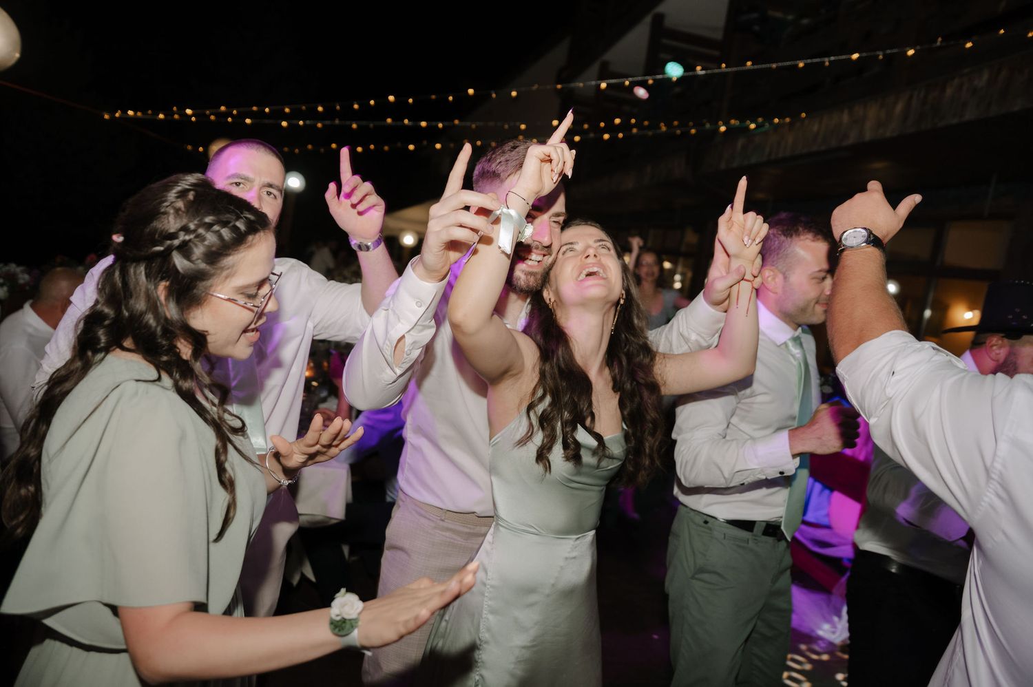 Wedding guests dancing and celebrating on the dance floor during a reception with festive lighting.