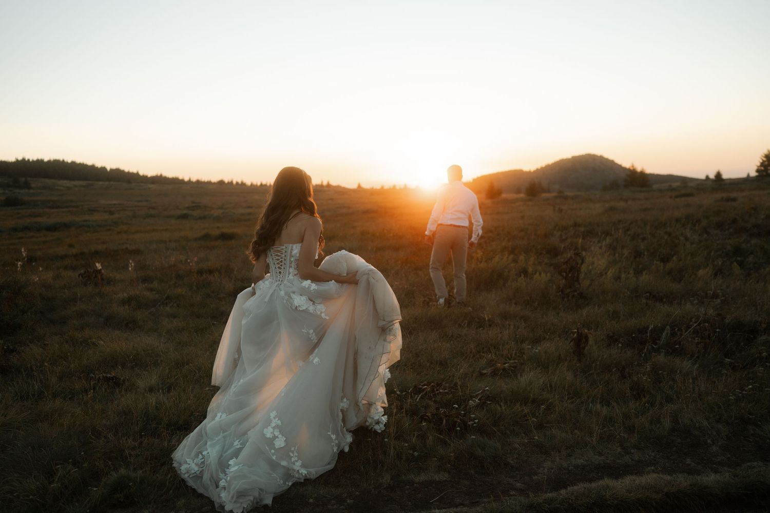 A couple walks through a mountain field at sunset with the bride's dress flowing in the breeze.