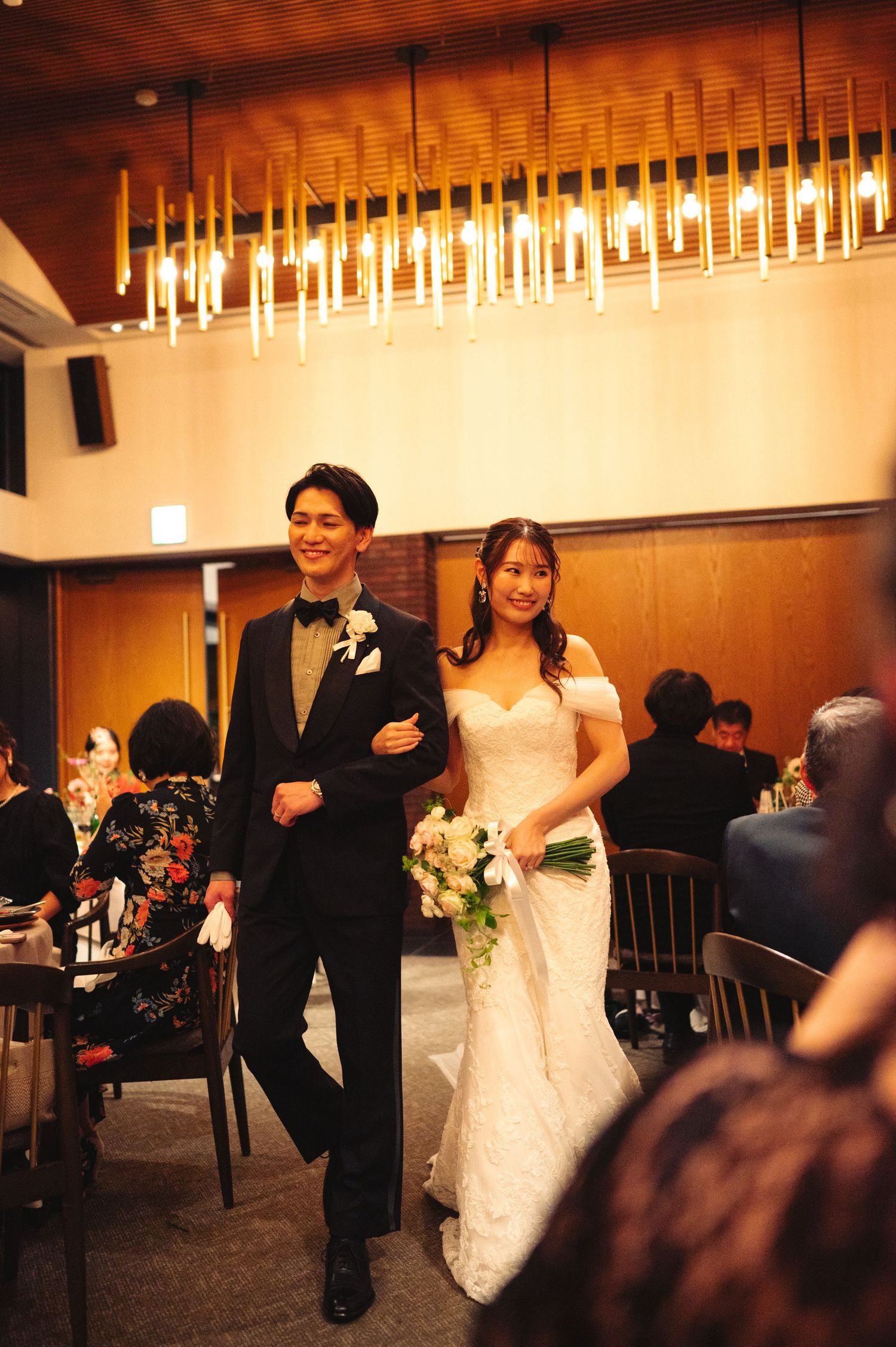A couple walks down the aisle at their wedding reception under elegant pendant lighting.