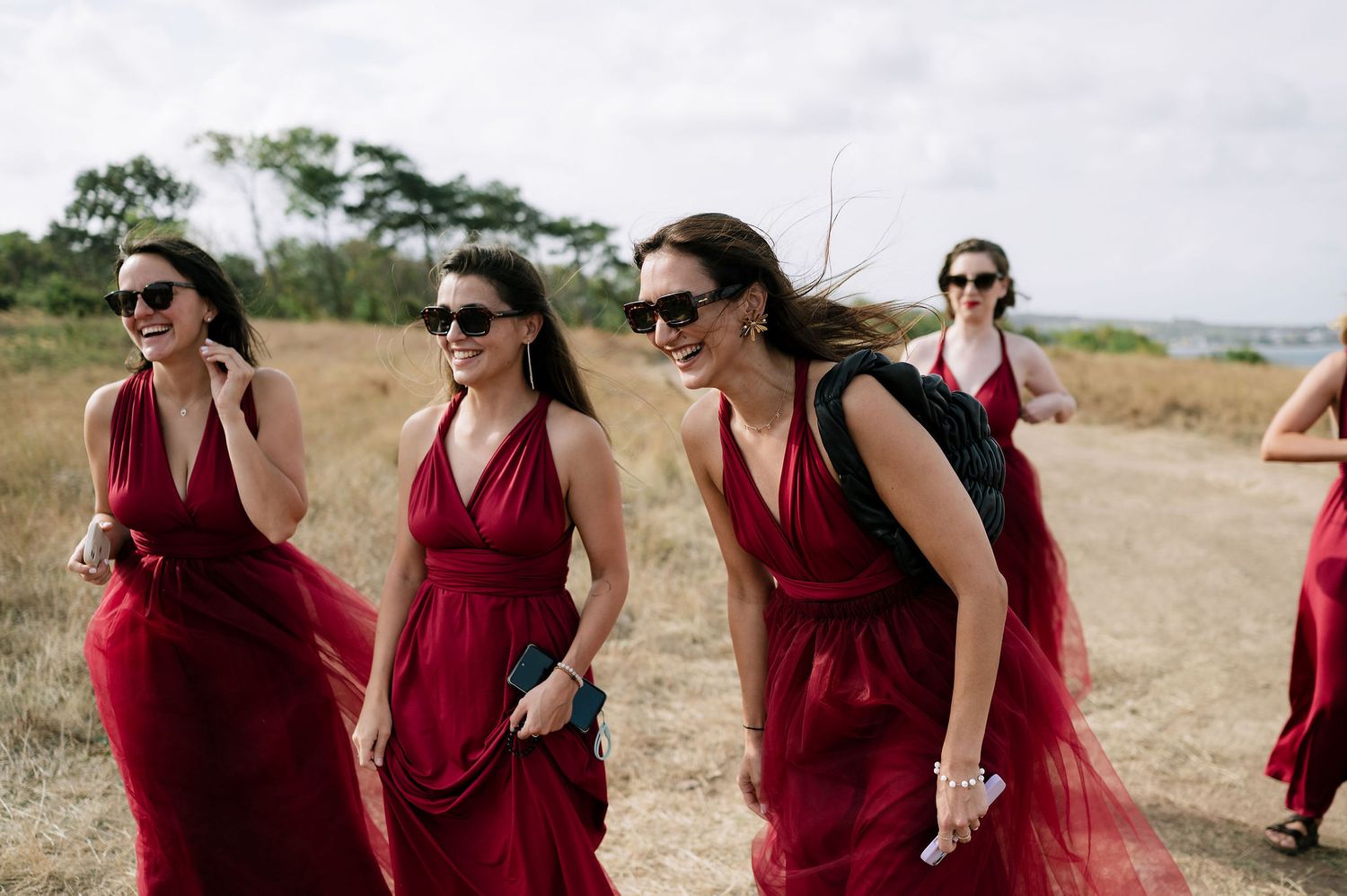Bridesmaids in flowing burgundy dresses walk together through a scenic outdoor field.