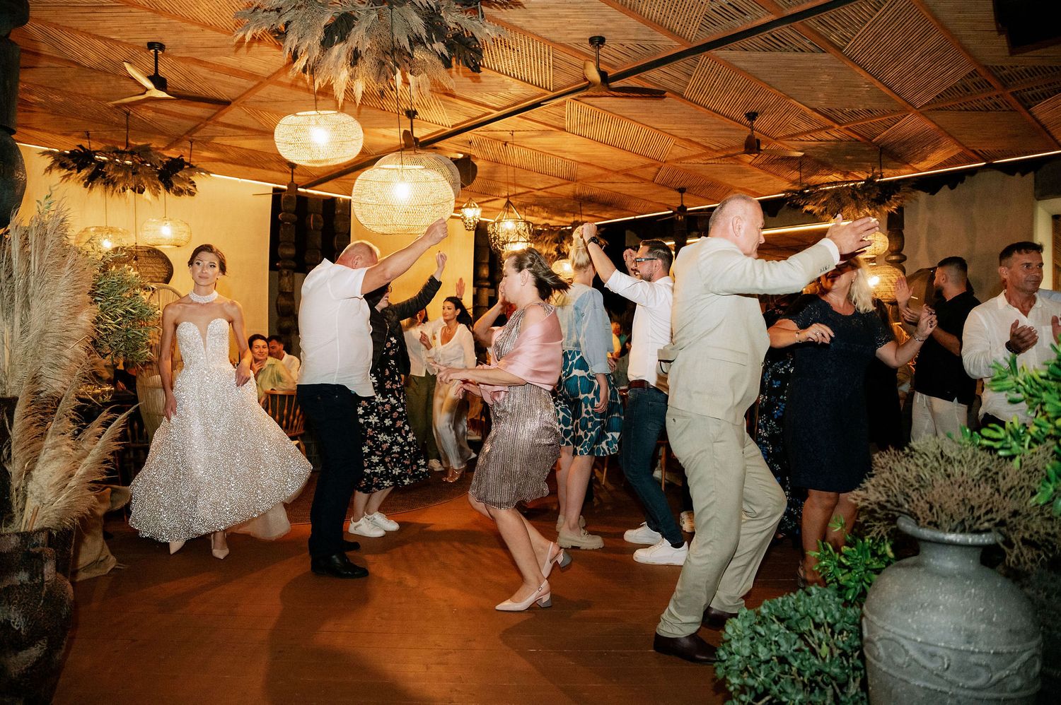 Wedding celebration with guests dancing in a rustic venue decorated with pampas grass and potted plants.