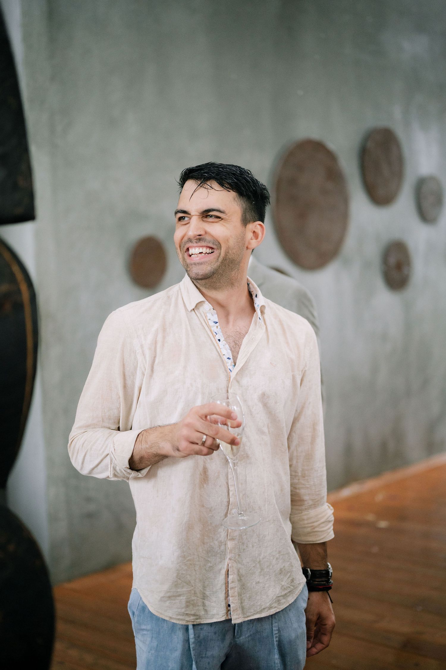 Person in beige linen shirt laughing while holding a drink.