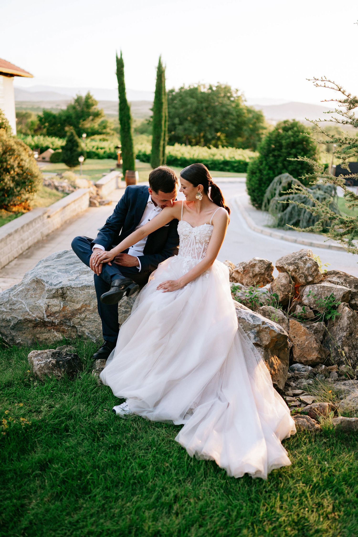 Bride in flowing white gown and groom share tender moments on landscaped garden rocks.