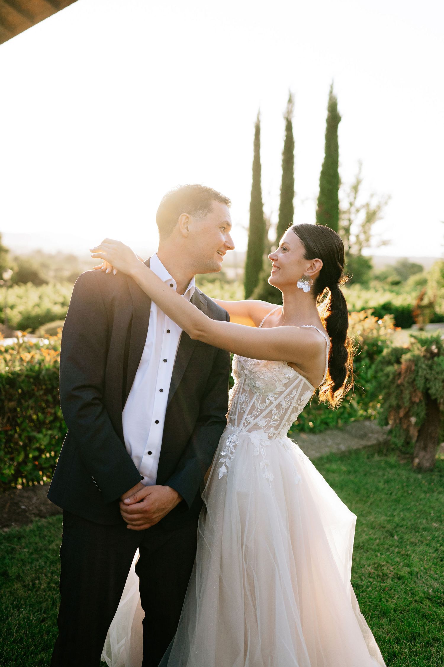 Wedding couple shares playful dance moves on a lawn with cypress trees during golden hour.