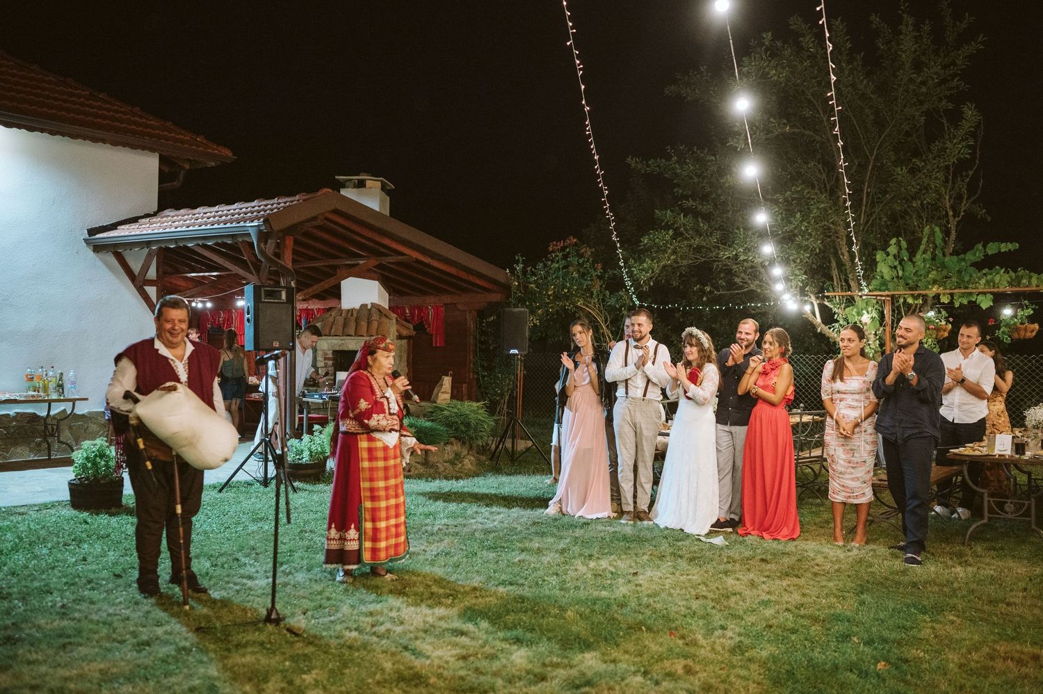 Wedding celebration guests gathered on lawn under string lights at dusk.