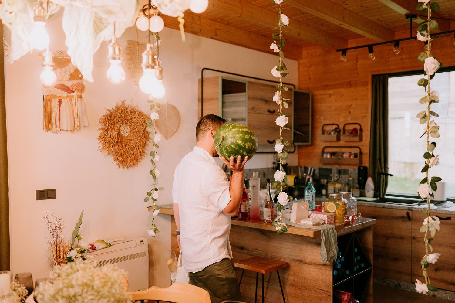 A person in white prepares drinks at a rustic wooden bar decorated with hanging lights.