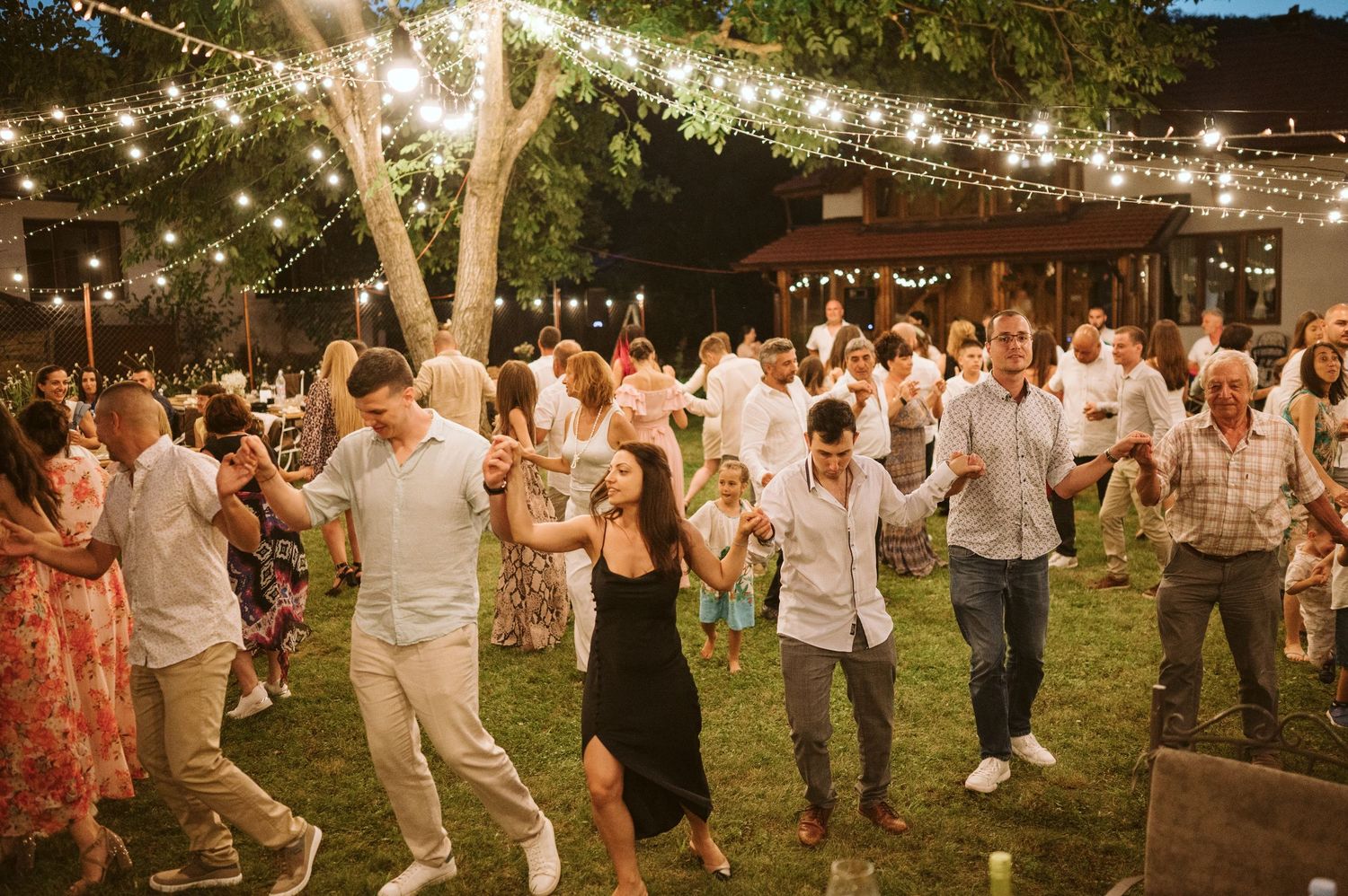 Large group of wedding guests dancing on a lawn under festive string lights at night.