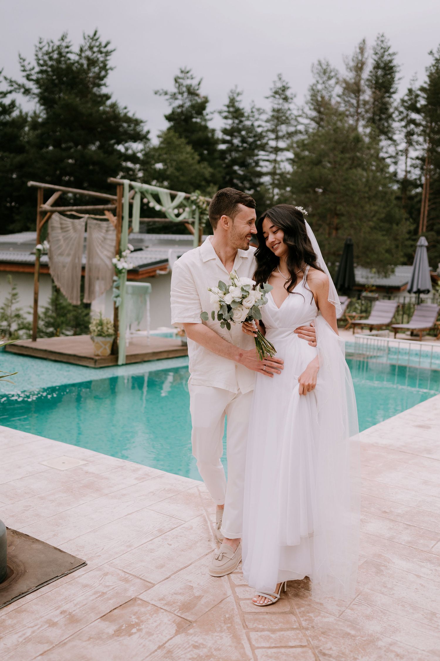 Wedding couple in all-white attire poses together near a resort swimming pool.