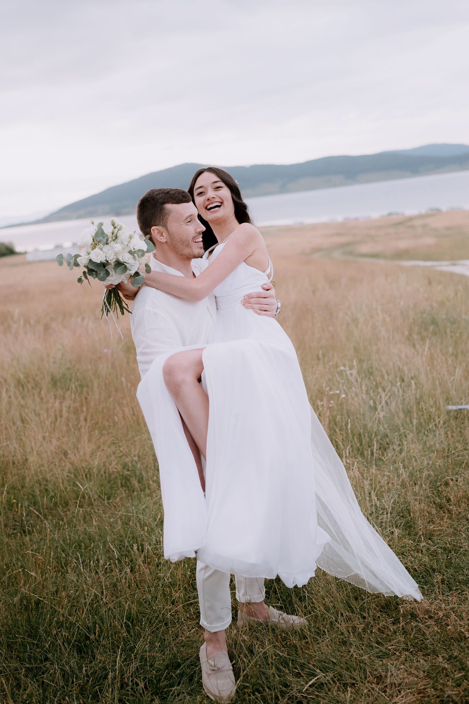 Joyful couple sharing playful moment in seaside field with white wedding attire.