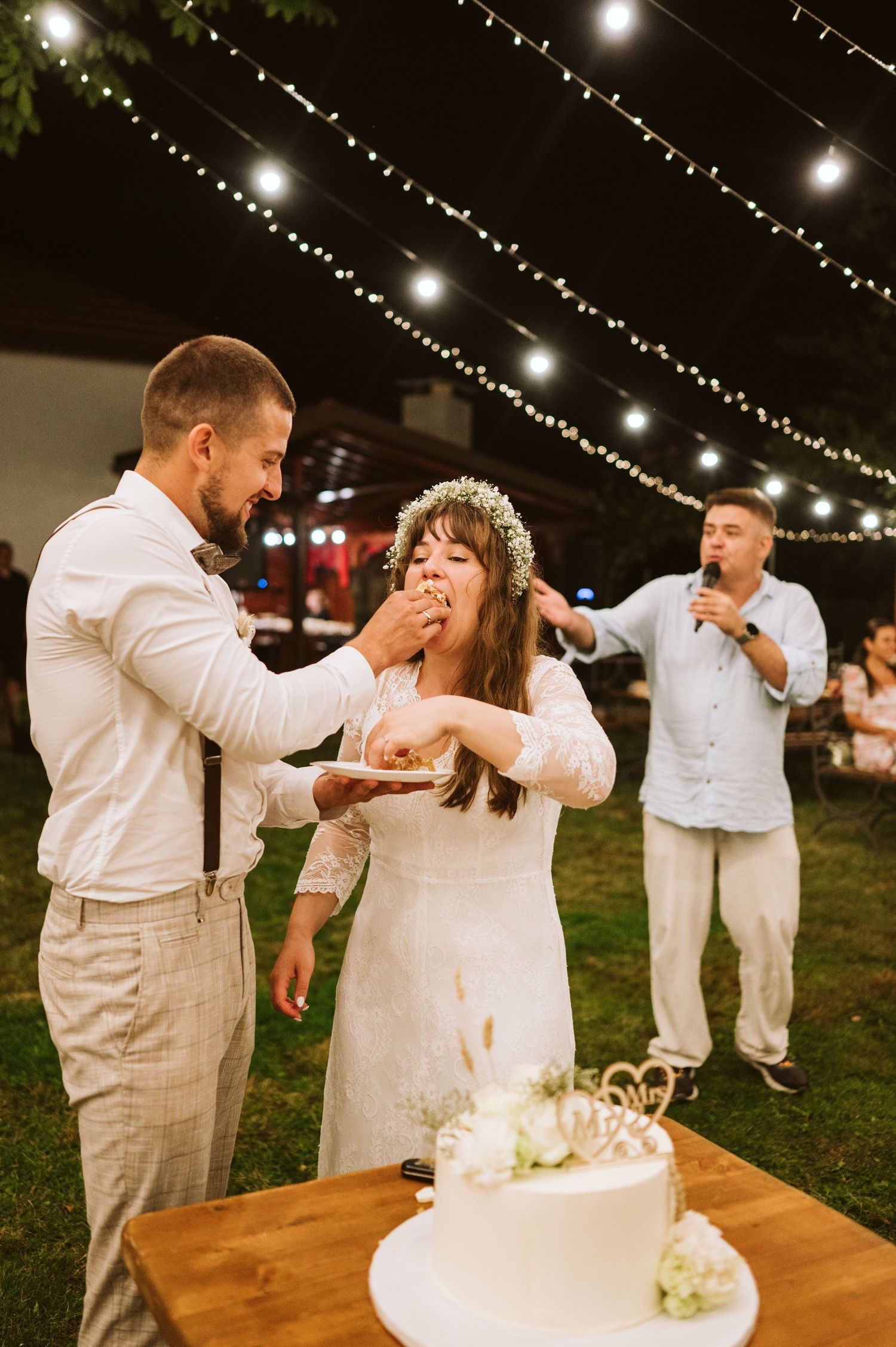 Newlyweds share a playful cake-cutting moment under string lights at an outdoor wedding reception.