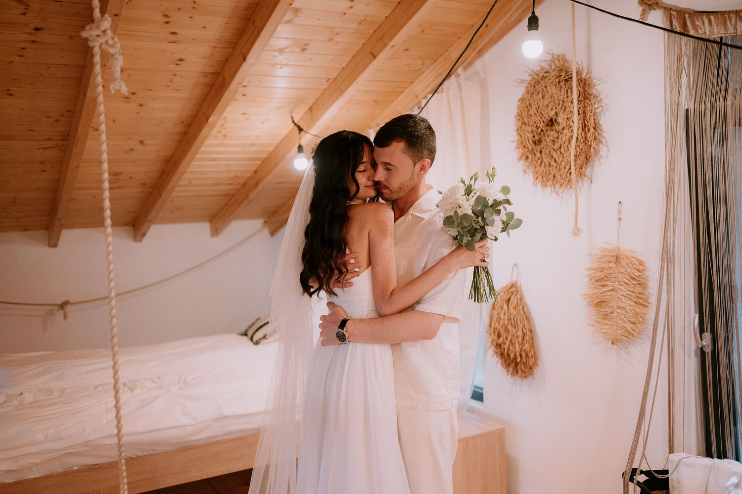 Artistic wedding scene in rustic attic with hanging decorations and warm lighting.