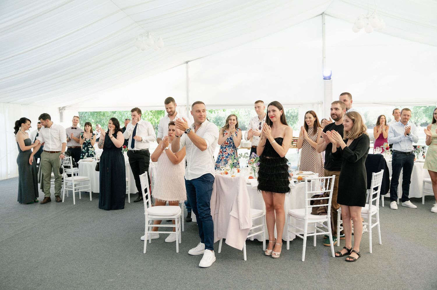 Wedding guests stand and applaud during reception celebration under white tent.
