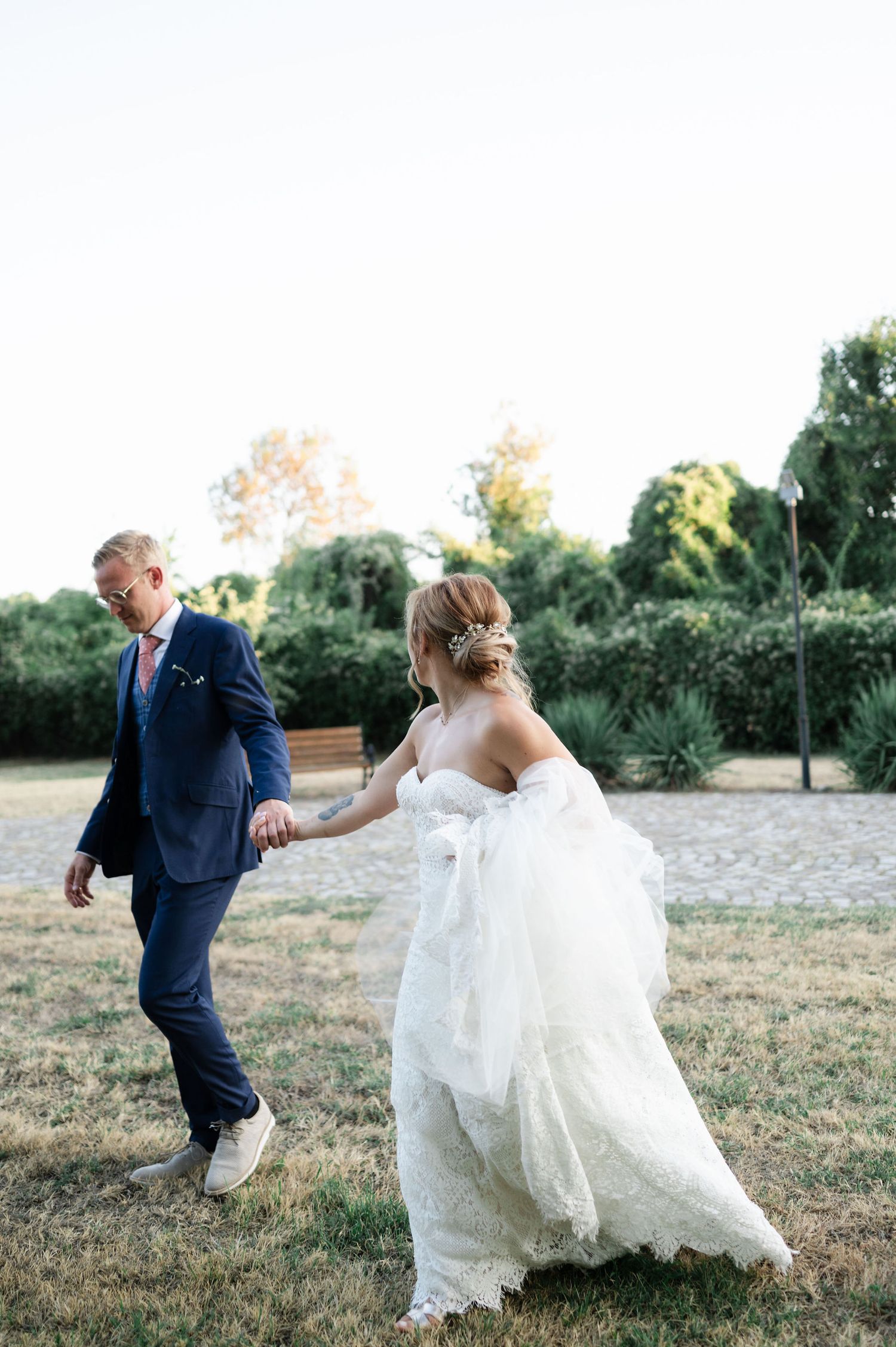 Wedding couple runs playfully through a grassy field in formal attire during golden hour.