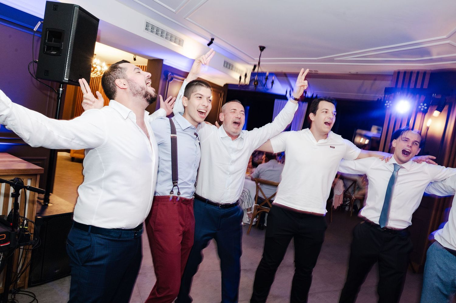 Large group dancing in formation with arms raised at an indoor wedding reception.