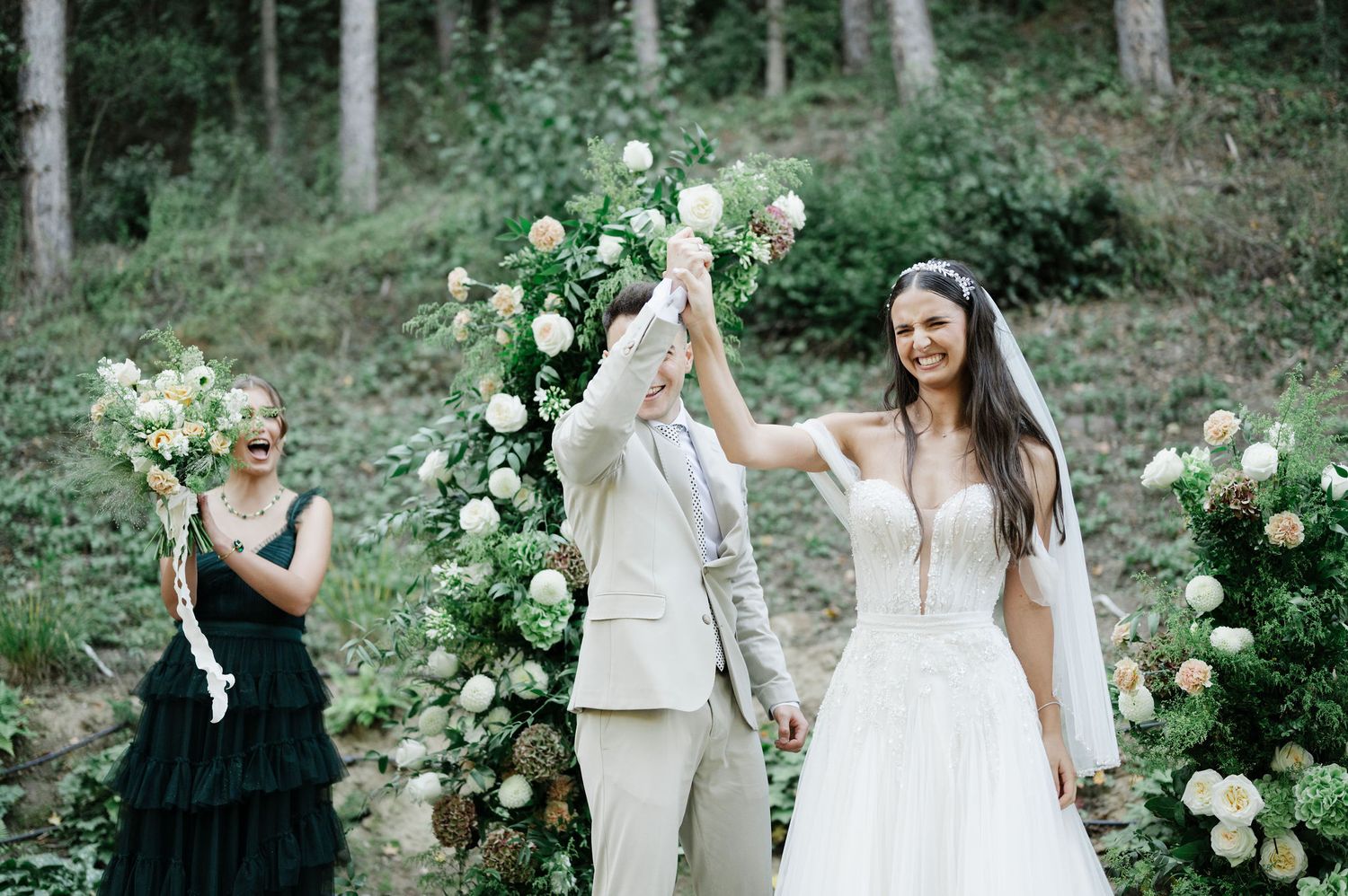Joyful celebration moment under floral arch at outdoor forest wedding.