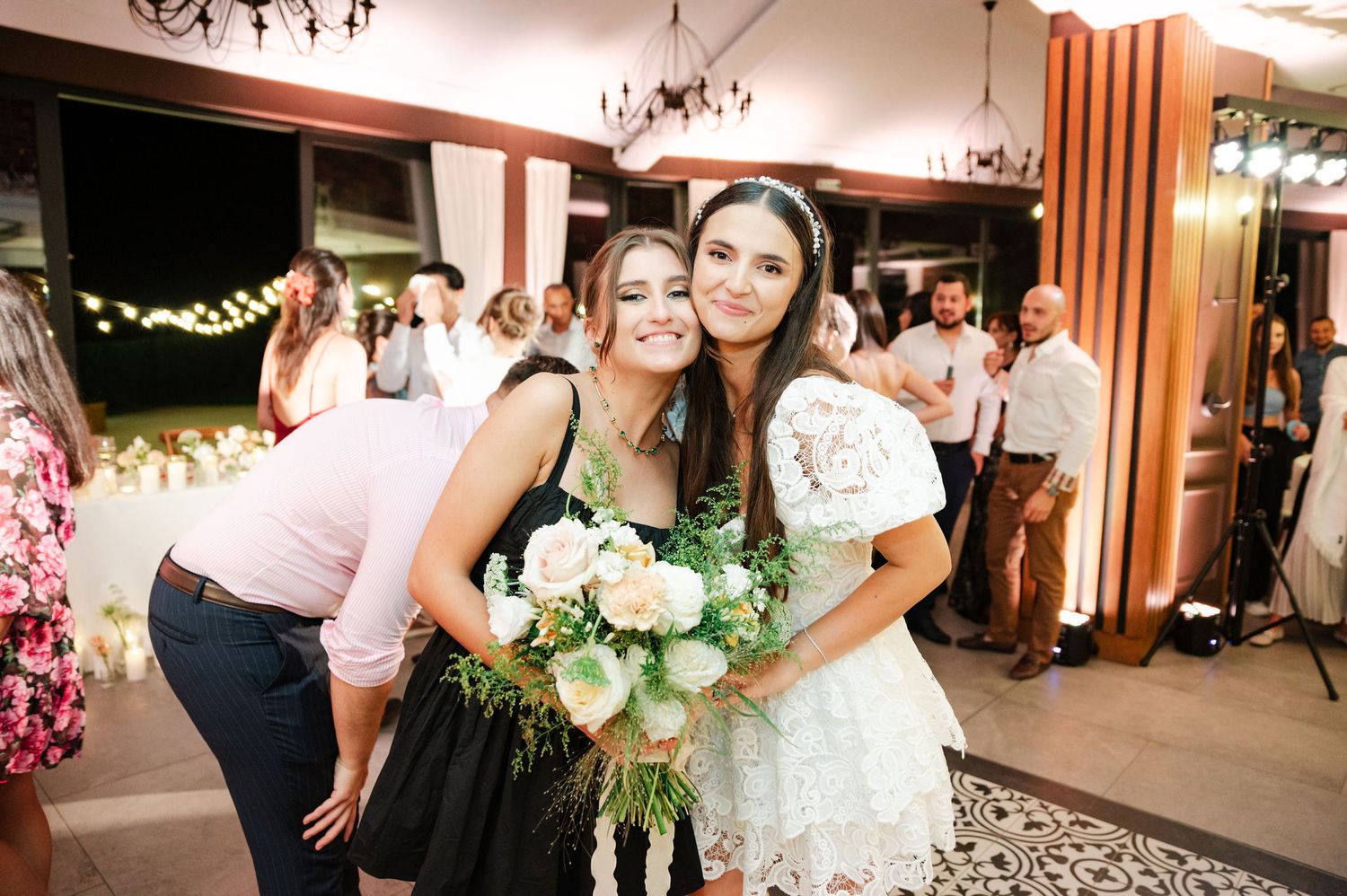 Two guests pose with a bouquet of white flowers at an indoor celebration.