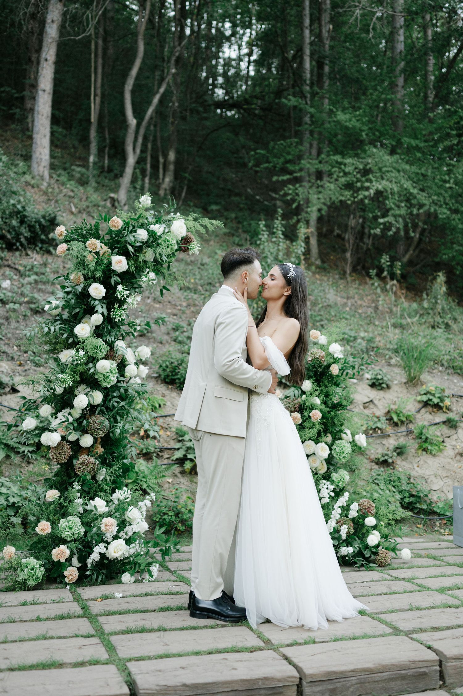 A romantic wedding moment captured in front of lush white floral arrangements in a forest setting.