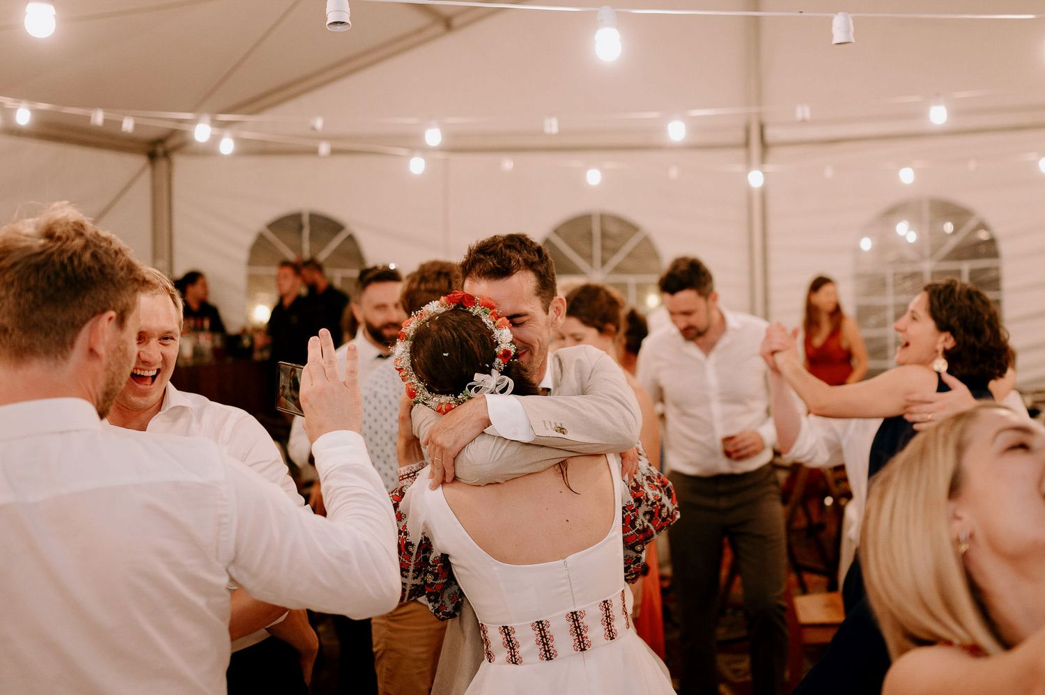Guests celebrating at a wedding reception with string lights overhead in tent venue.