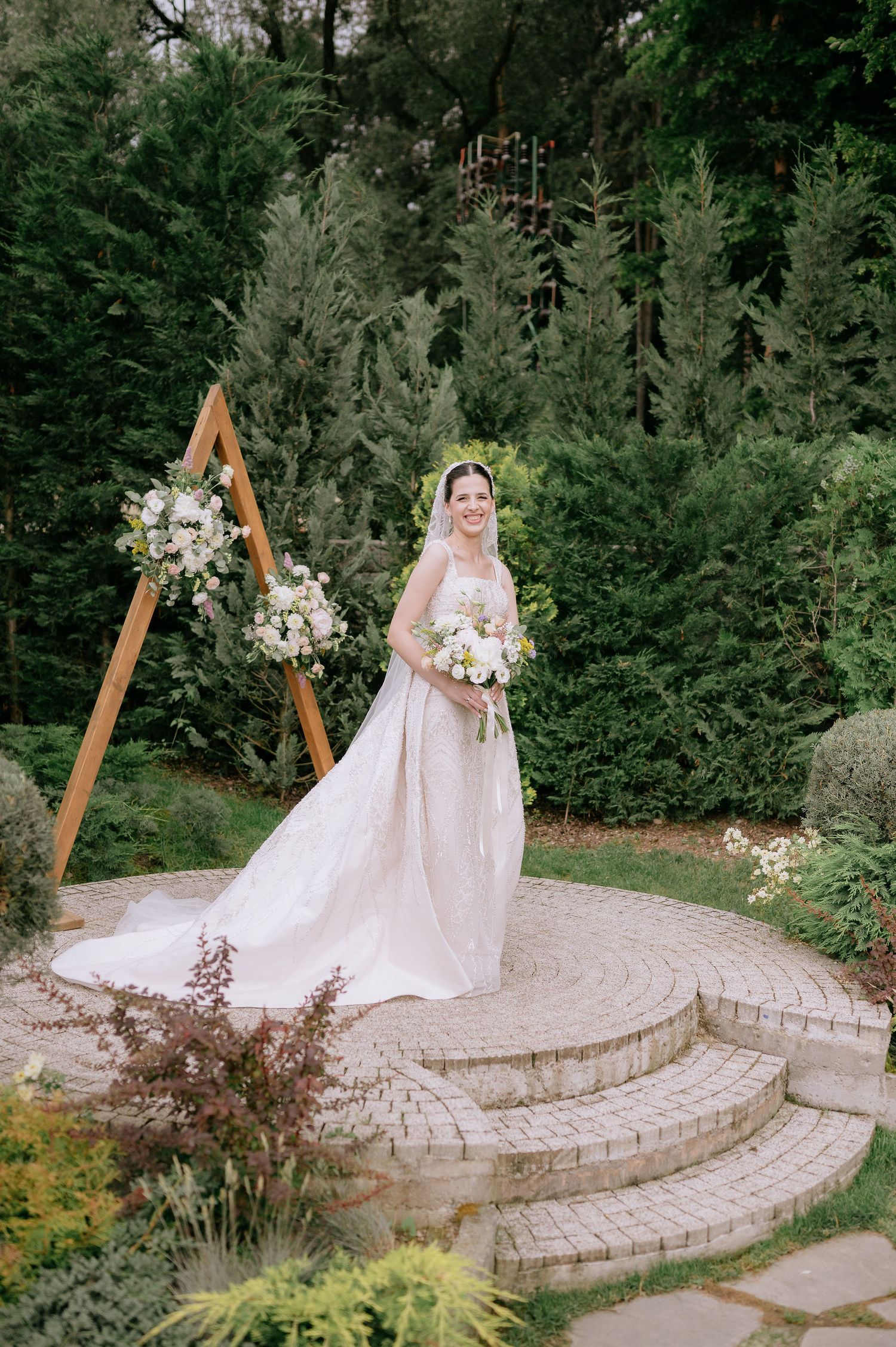 Wedding dress and bouquet photographed on tiered stone patio with wooden ceremony arch.