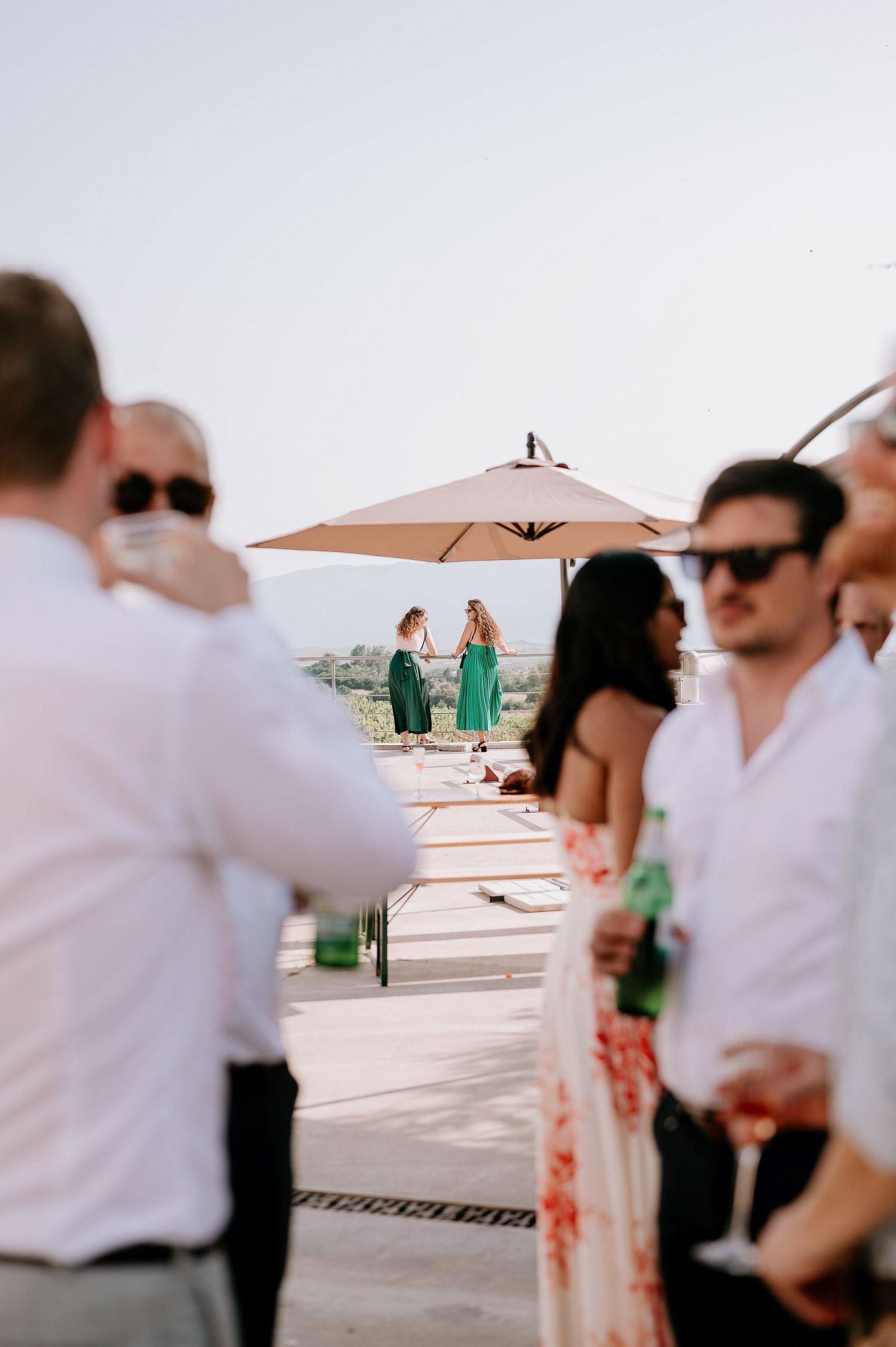 Wedding guests mingling on outdoor terrace with umbrellas and scenic views.
