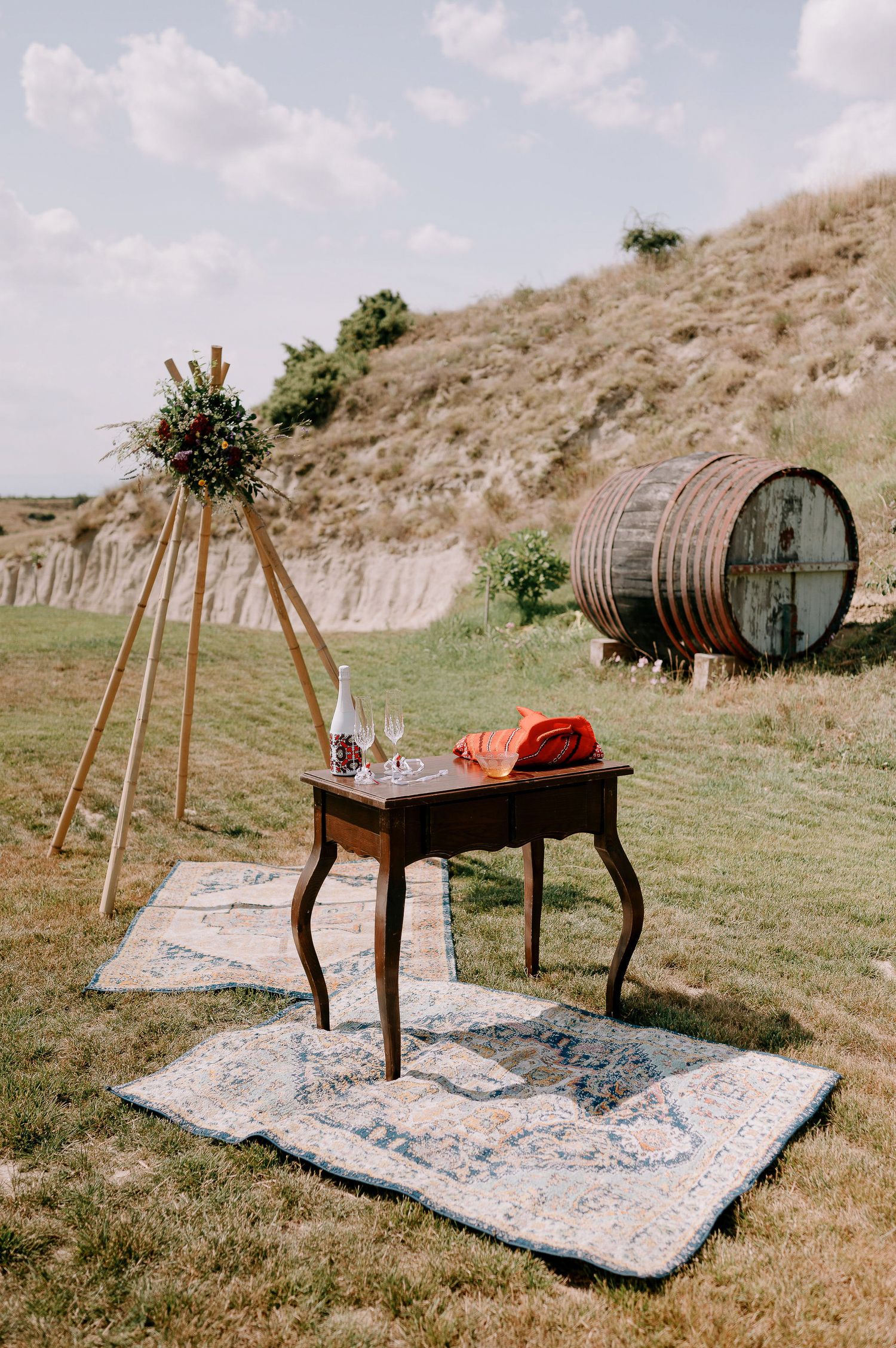 Rustic wooden table with wine setup and large barrel in background on grassy hillside.