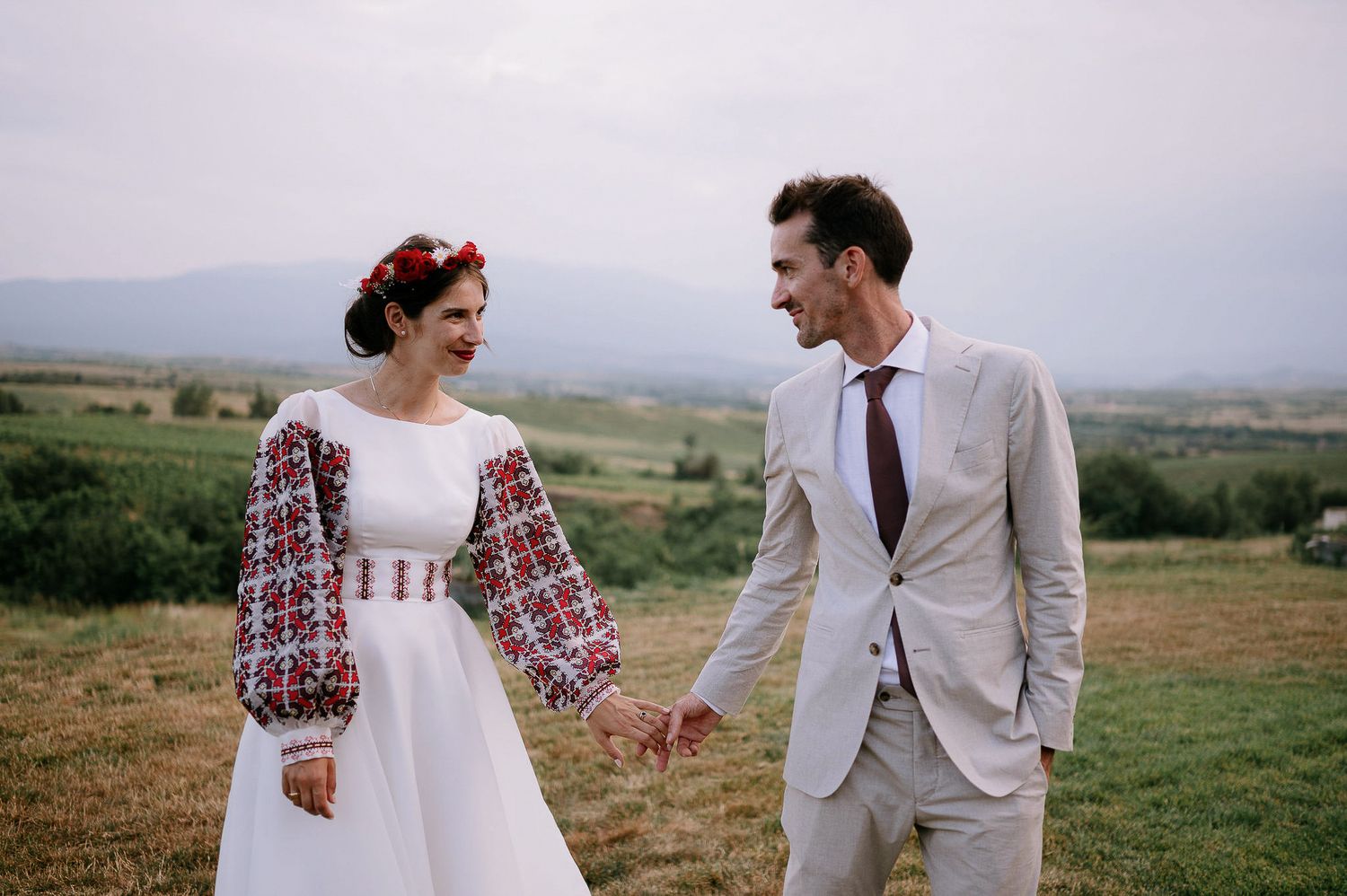 A couple in traditional wedding attire laughs together on a grassy hilltop with mountain views.