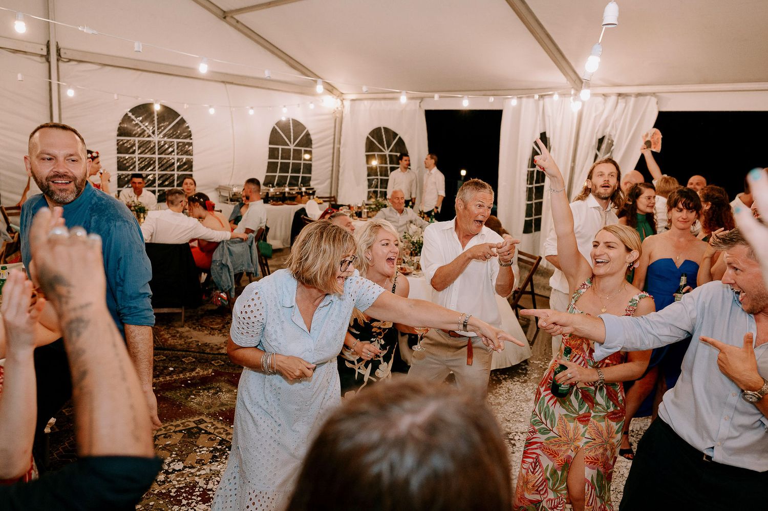 Festive wedding reception with guests dancing under string lights in an outdoor tent.