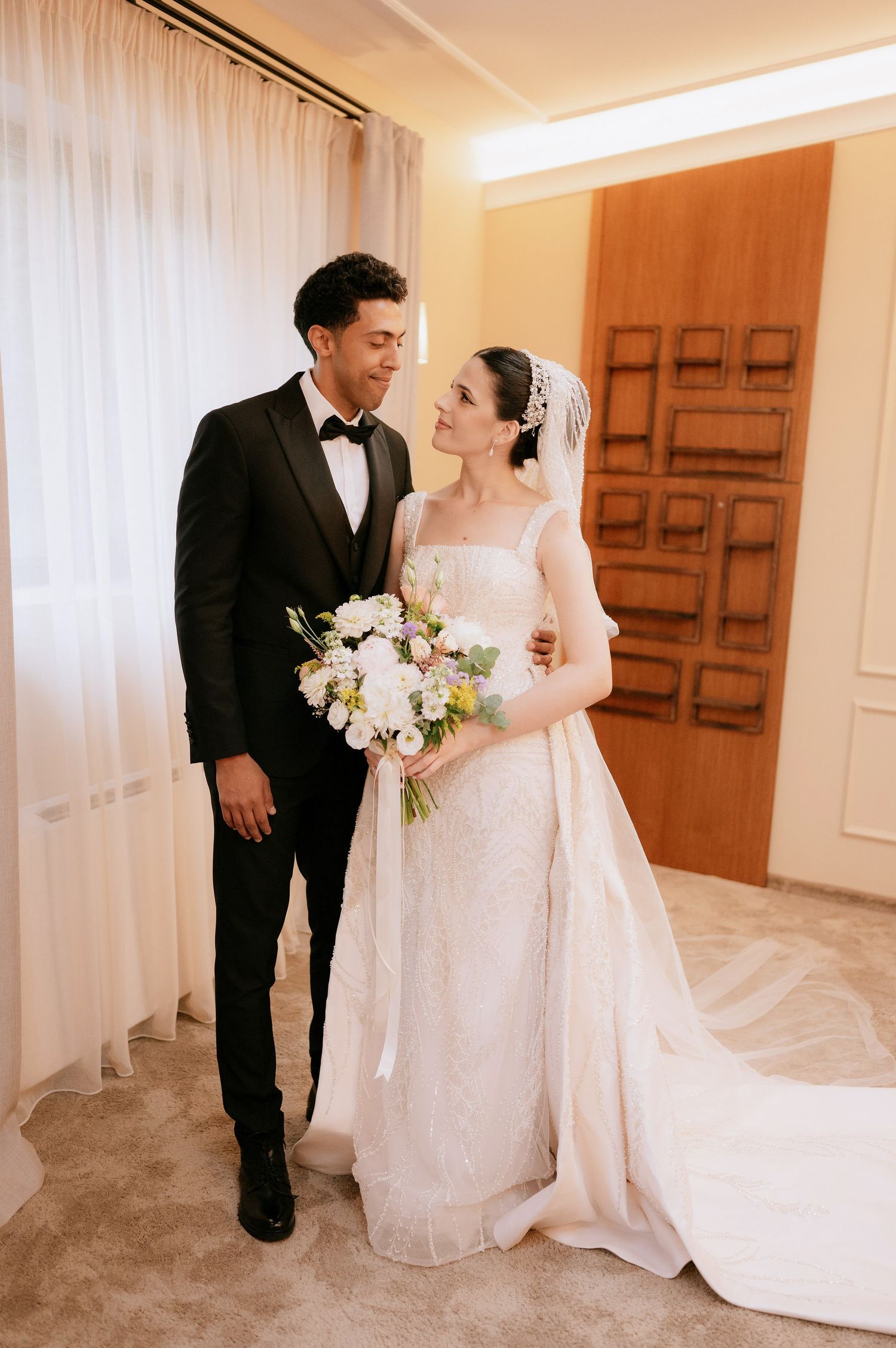 A couple in formal wedding attire stands together in a bright indoor setting with white decor.