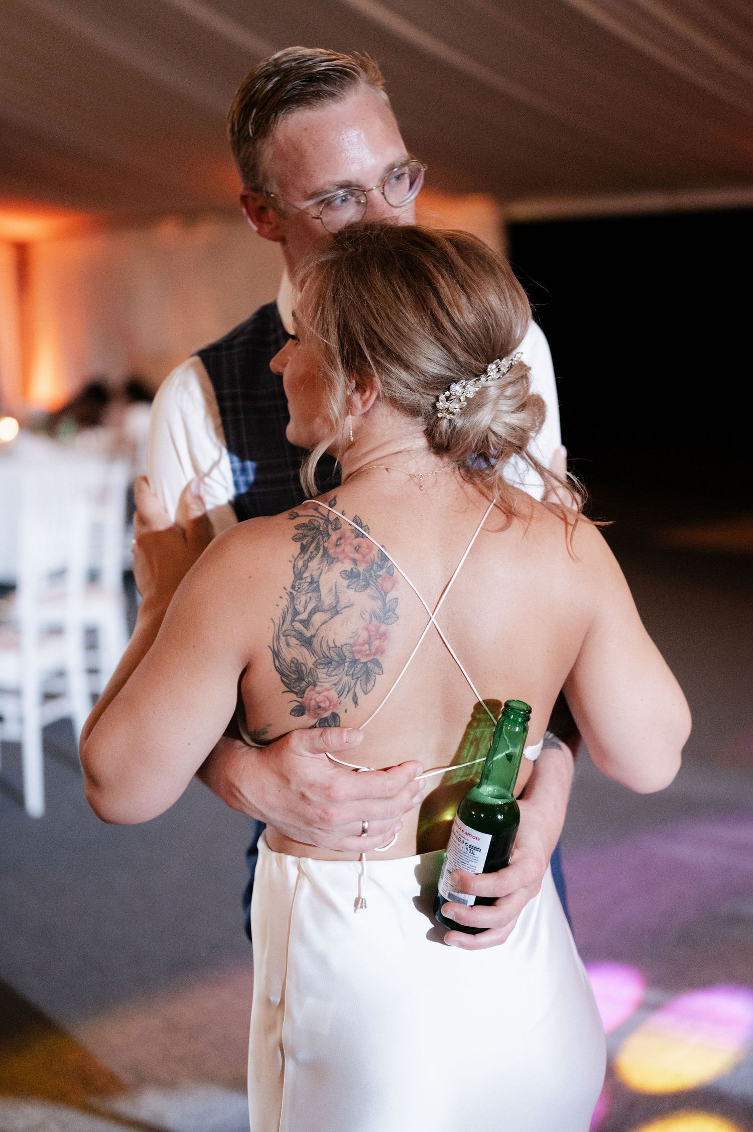 A bride with visible tattoos holds a drink while dancing at her wedding reception.