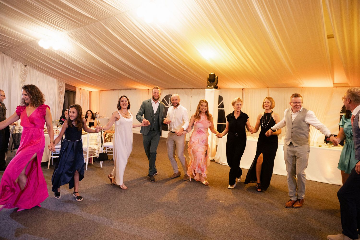 Wedding guests dancing and celebrating on the dance floor at an indoor reception venue with warm lighting.