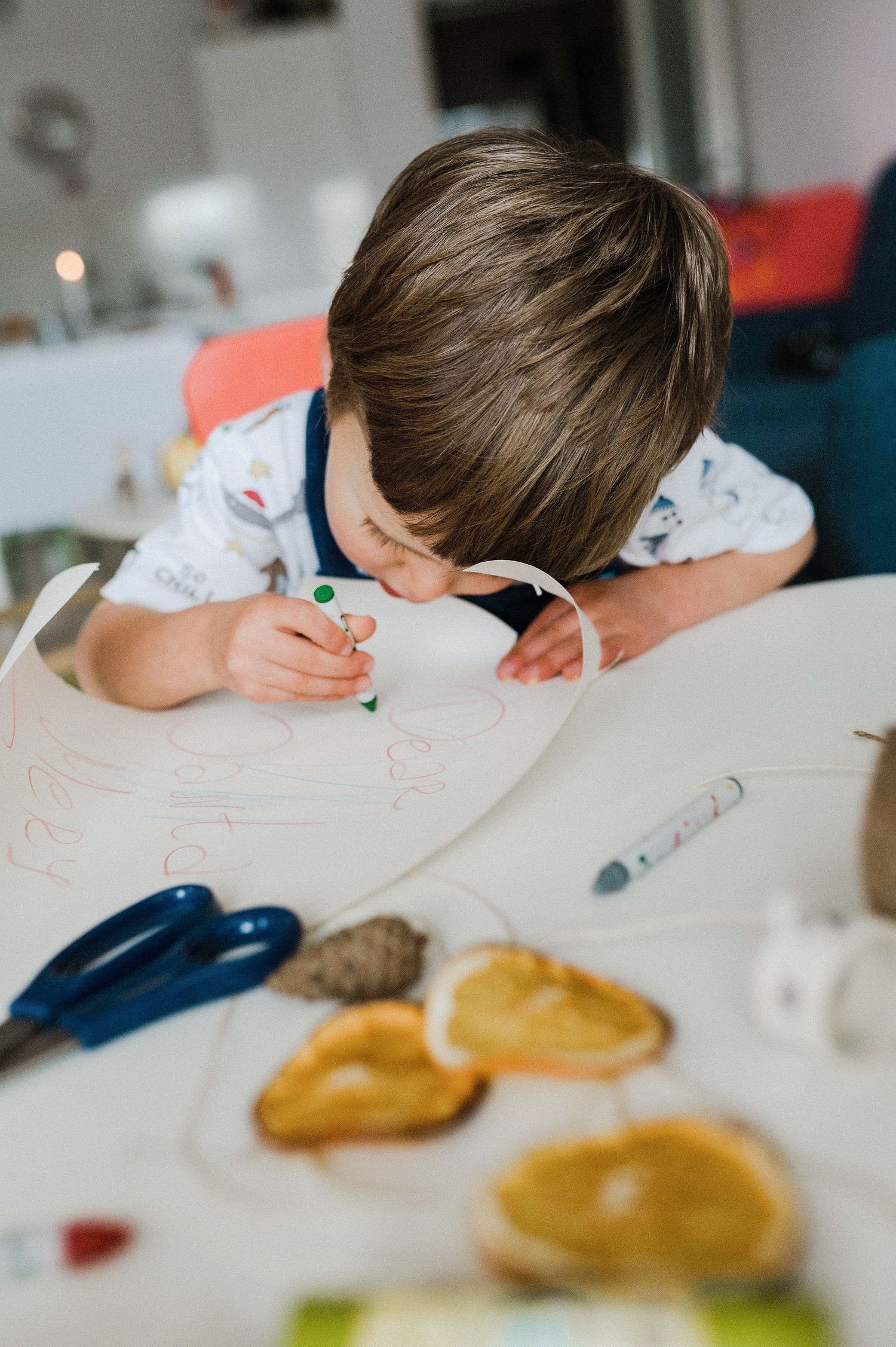 Child drawing or writing at a white table with food or snacks visible in the foreground.