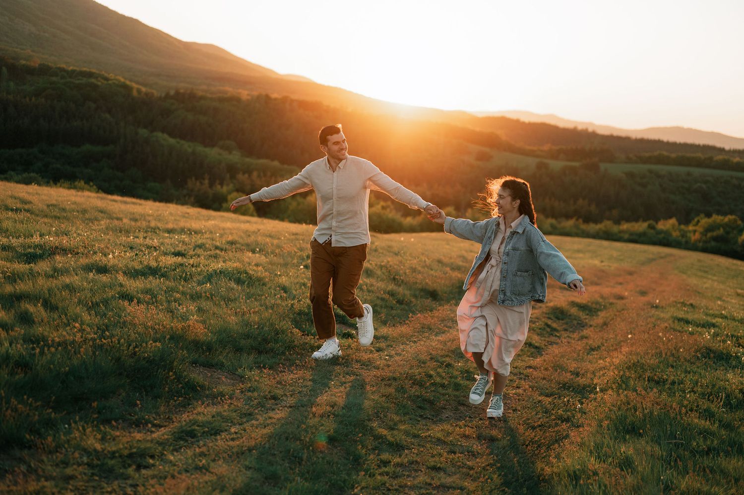 A couple runs playfully through a sunlit meadow at golden hour, creating a romantic silhouette against the landscape.