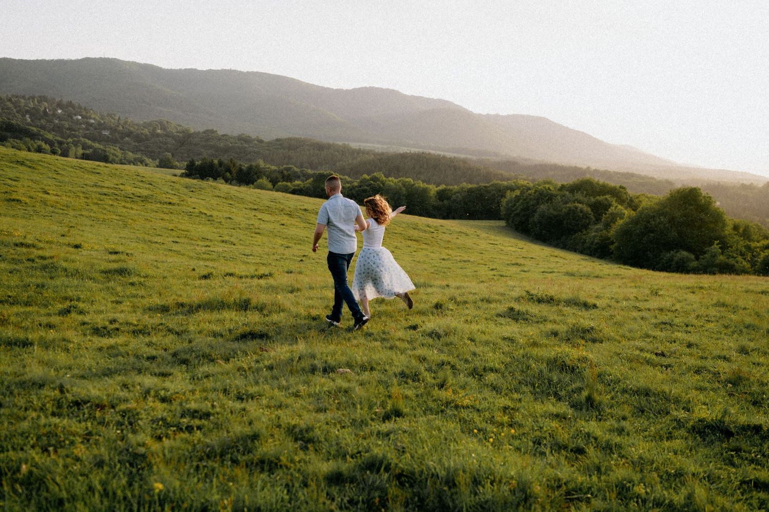 Two people dance joyfully in a hilltop meadow with mountains and sunset in the background.