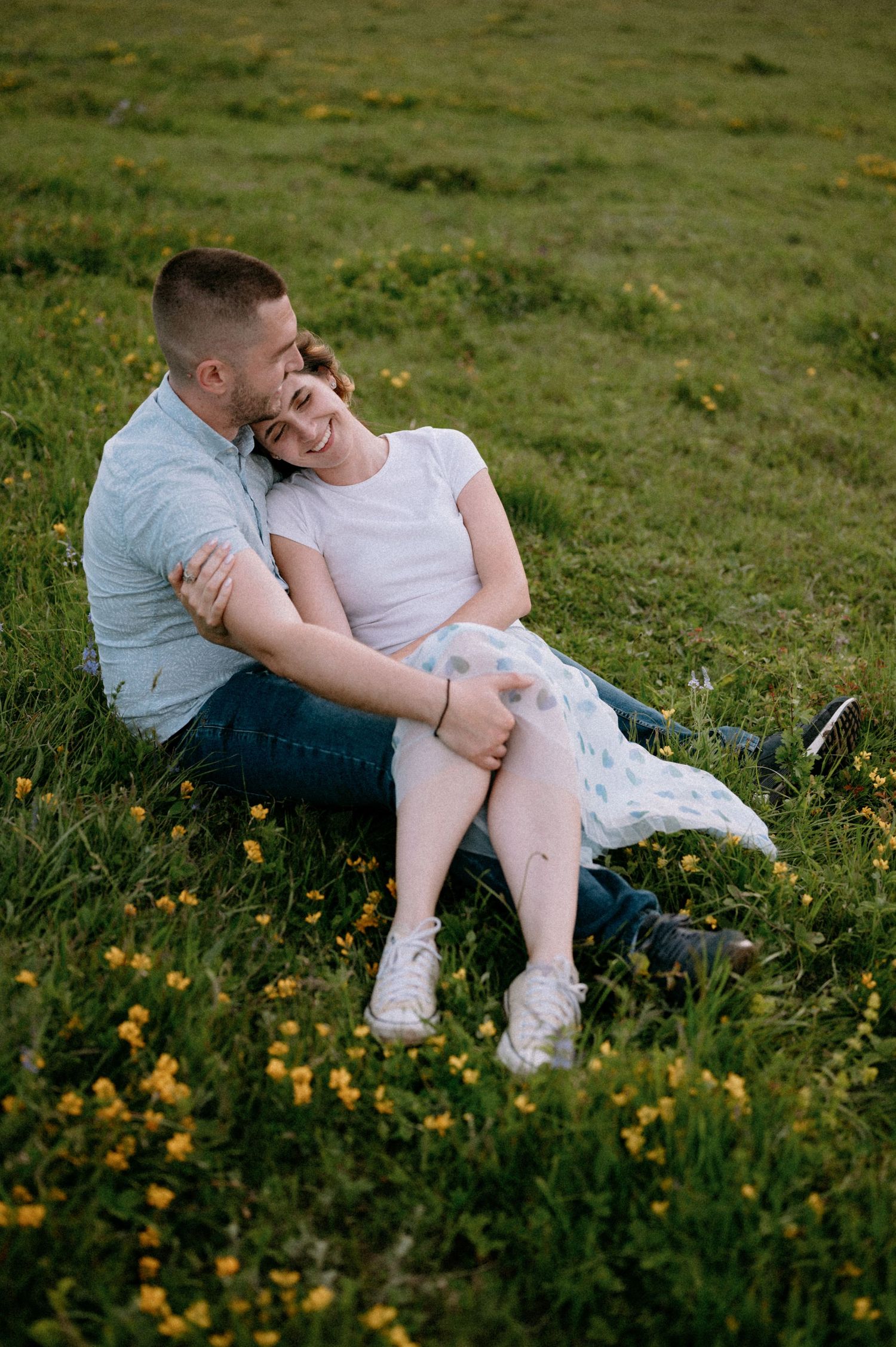 Couple relaxes together among yellow wildflowers on grassy mountainside.