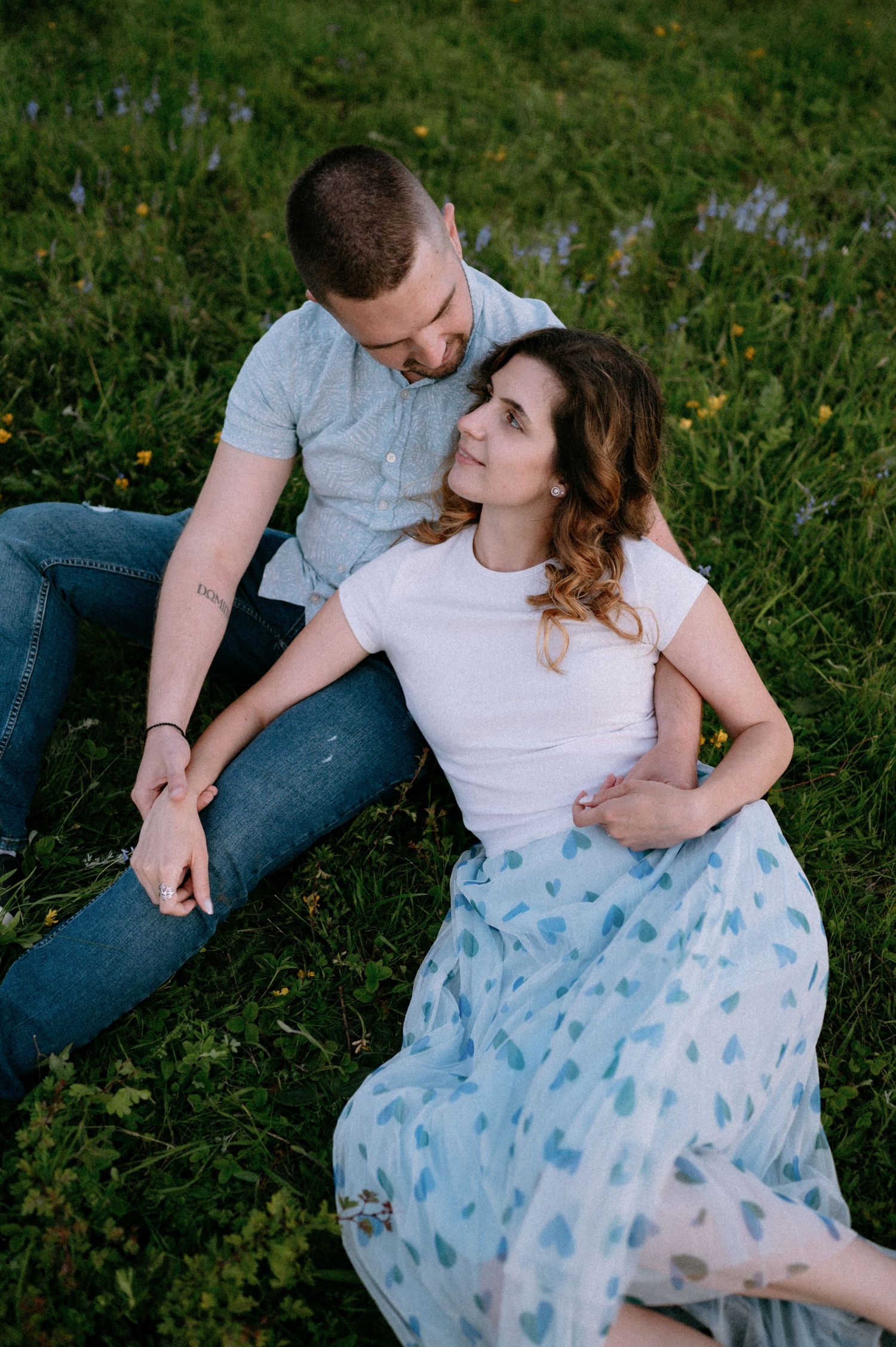 Two people share a tender moment while sitting in tall grass and wildflowers.