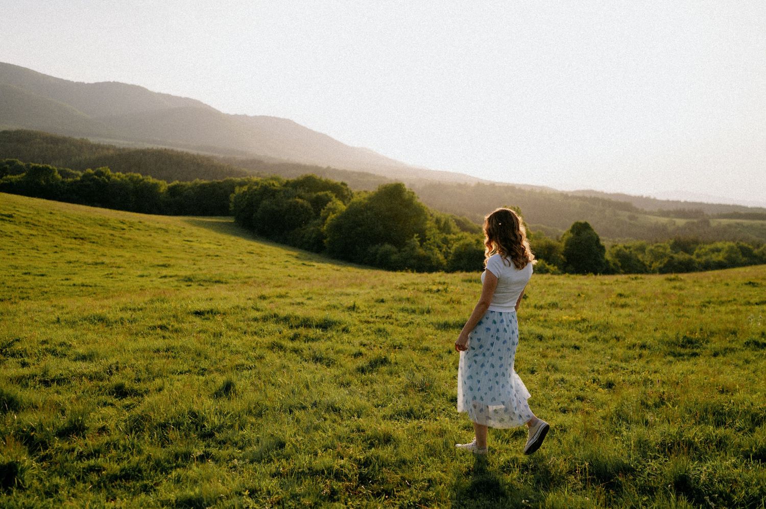 Figure in a flowing dress moves elegantly across a green hillside landscape.