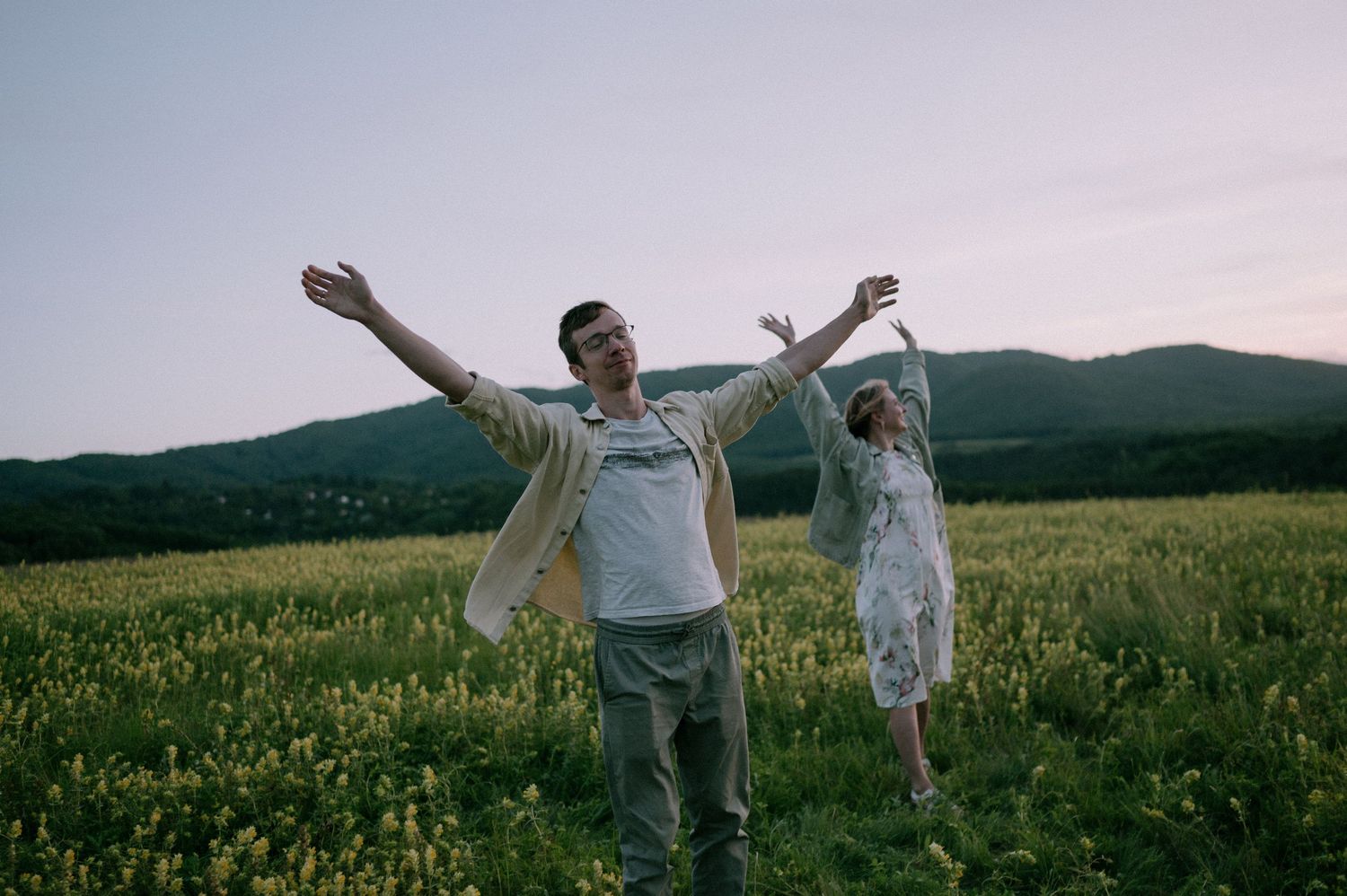 Two people celebrate with arms outstretched in a hilltop meadow during sunset.