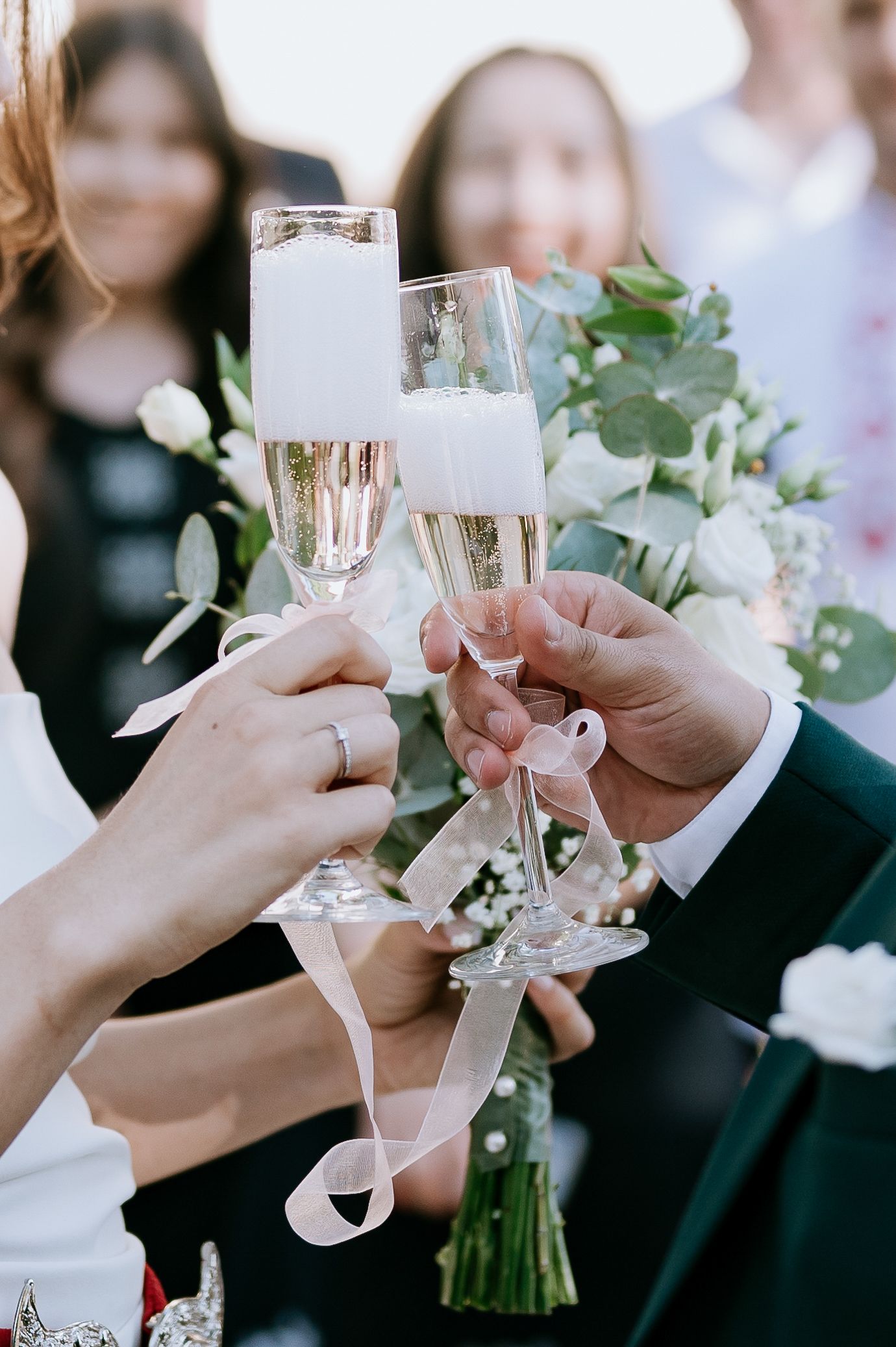 Champagne glasses clink in celebration with white ribbons and eucalyptus bouquet visible.