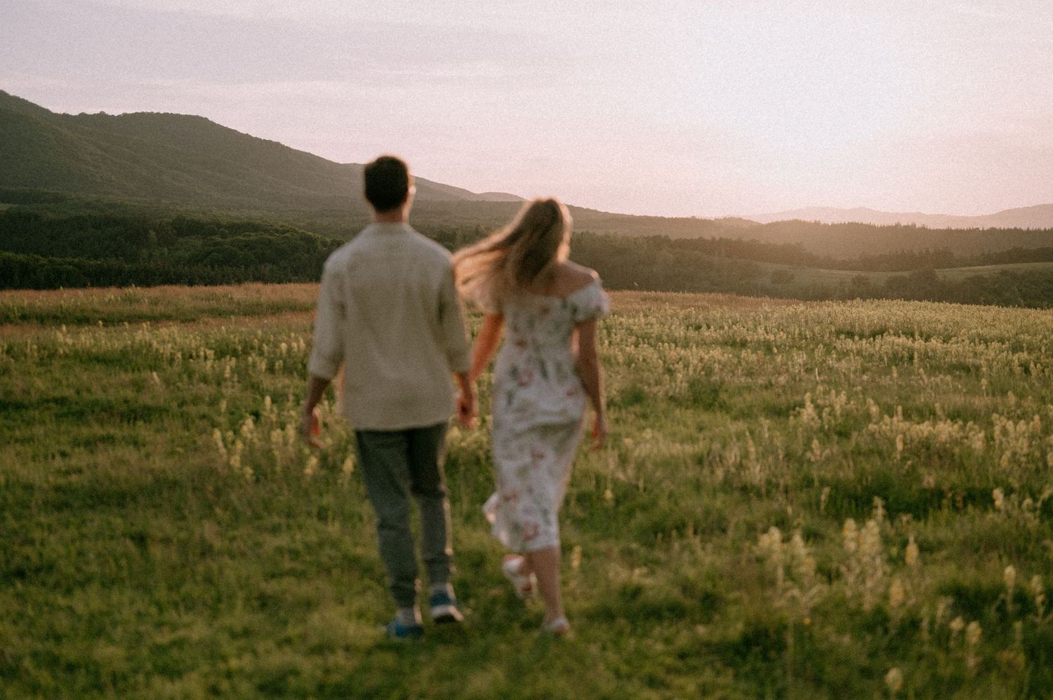A couple walks hand in hand through a scenic meadow at dusk with mountains in the background.