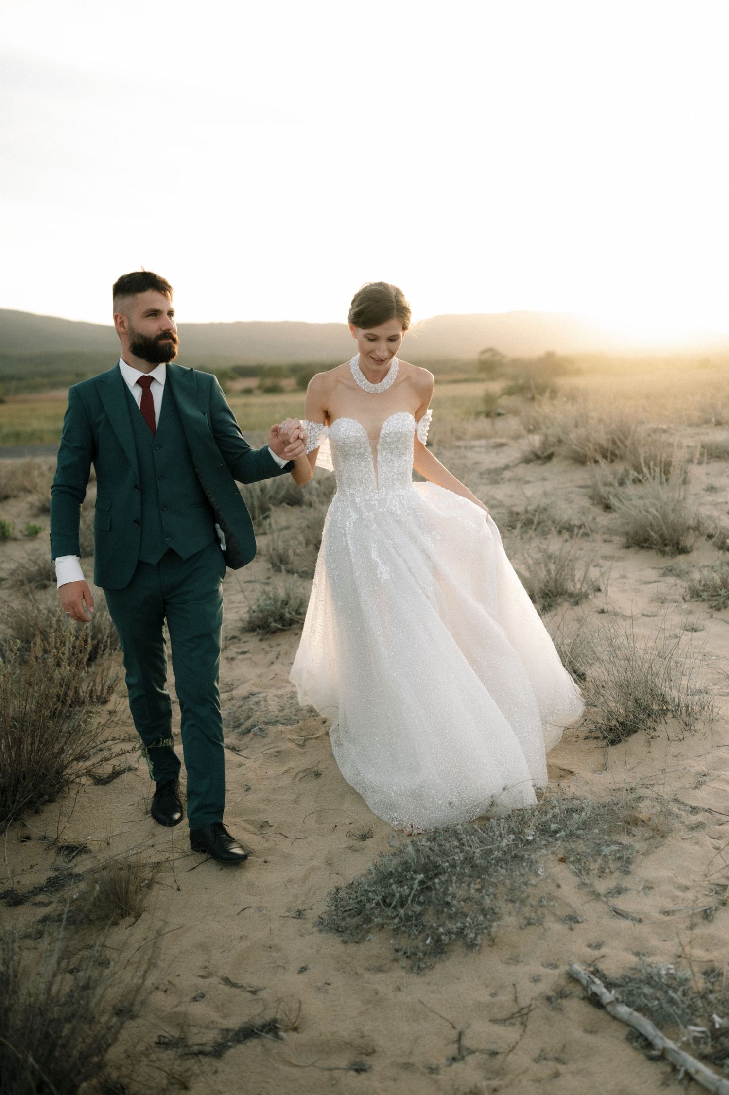 Bride and groom walk hand-in-hand through desert sand dunes at golden sunset, she in a flowing white gown.
